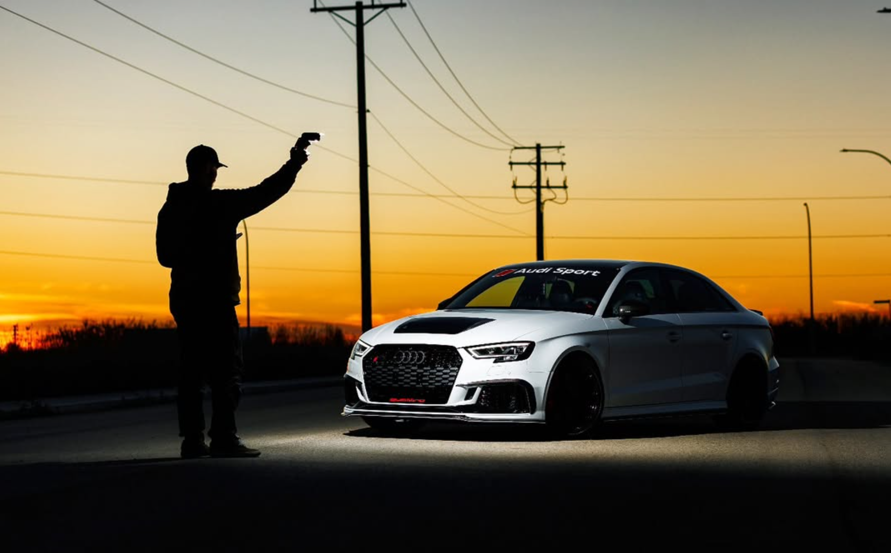 A silhouette of a man holding a gun pointed at a white Audi Sport car parked on the road at sunset, with power lines visible in the background.