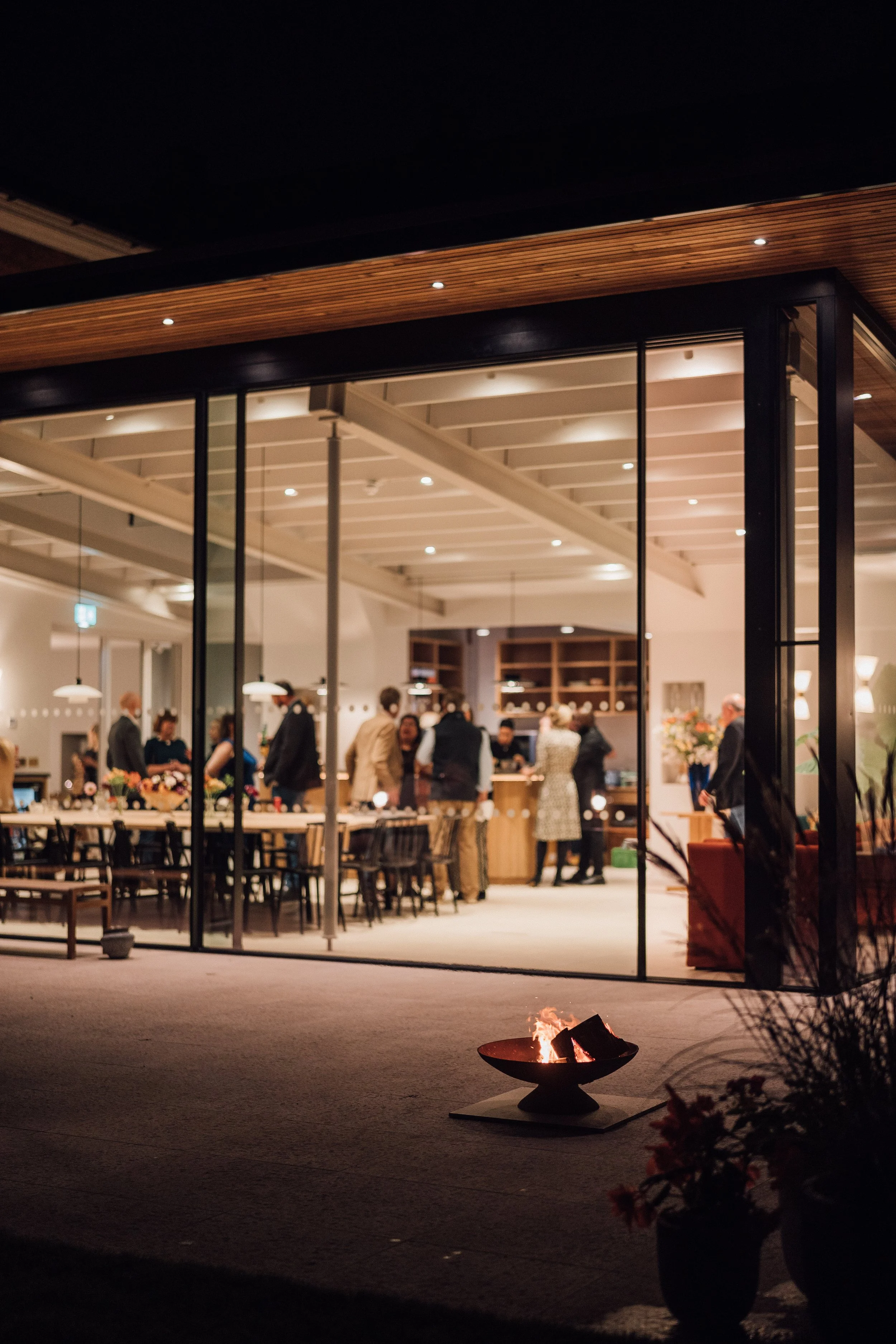 People inside a well-lit restaurant seen through large glass windows at night, with a visible flame fire on the patio outside.