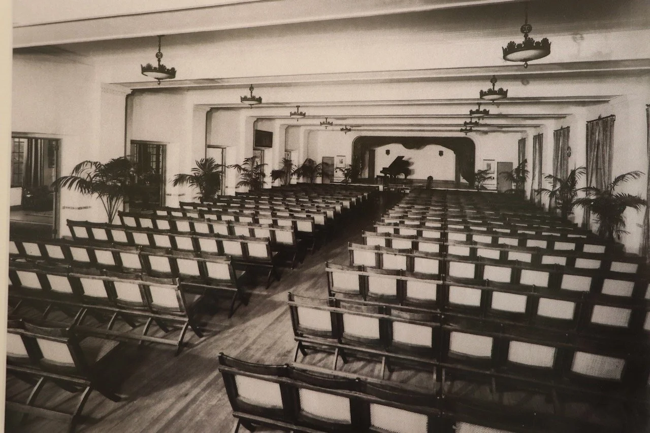 A vintage black-and-white photograph of an empty auditorium with rows of chairs, potted plants along the sides, and a stage with a grand piano at the front.