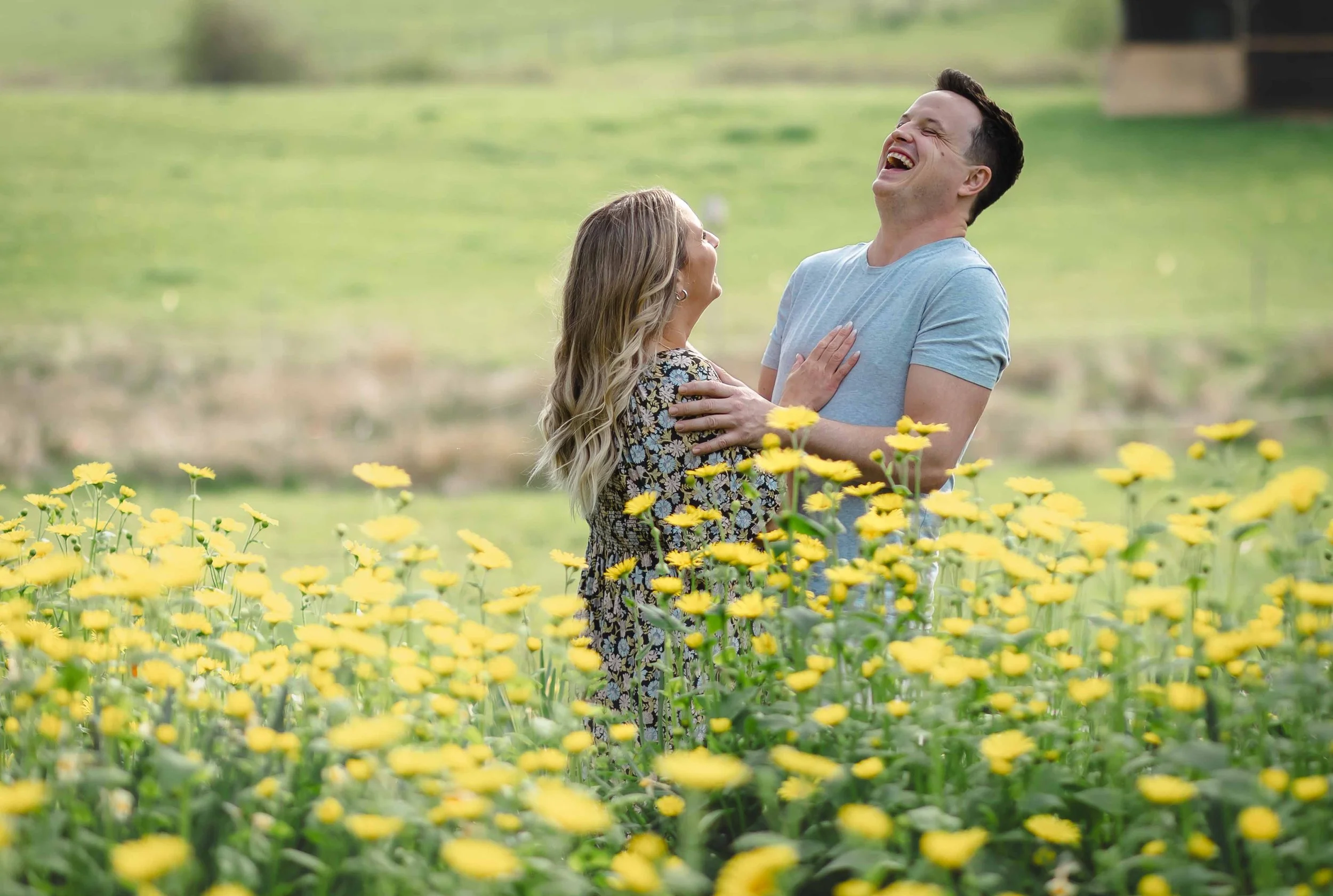 A man and woman laughing and embracing each other in a field of yellow flowers with a lush green background.