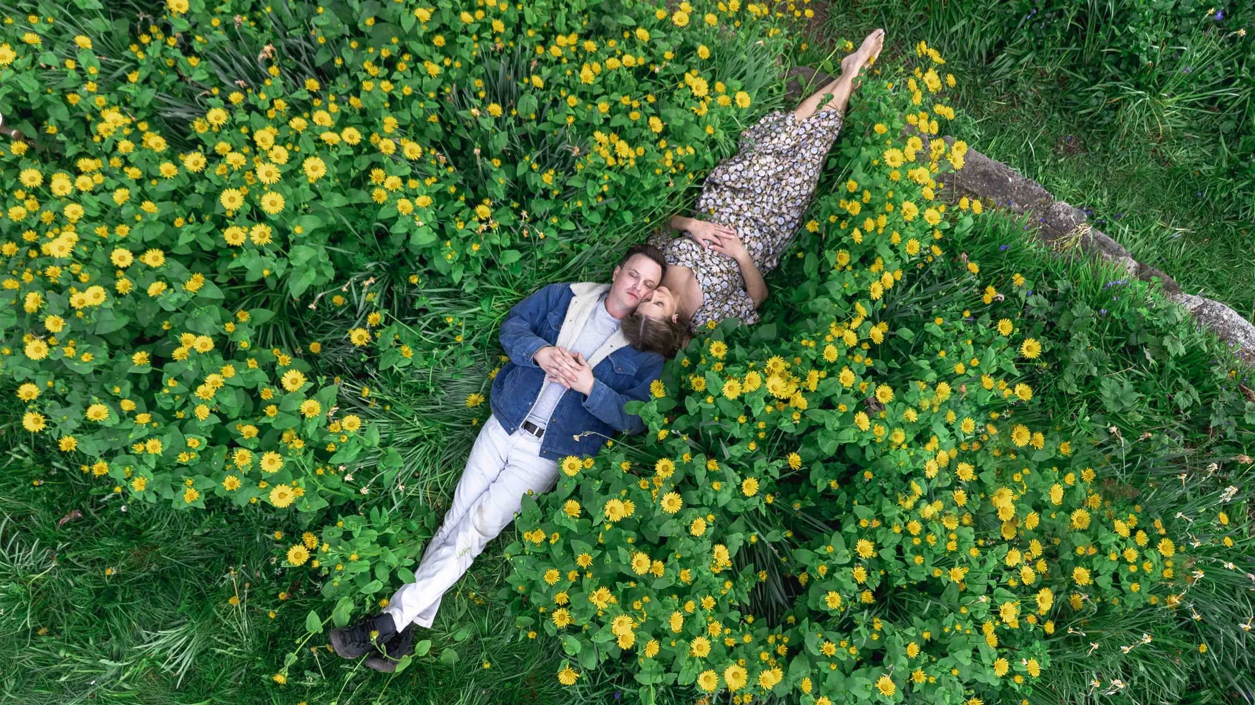 A couple lies on the grass among yellow flowers outdoors, with the woman wearing a floral dress and the man in a denim jacket and light-colored pants, both resting with closed eyes.