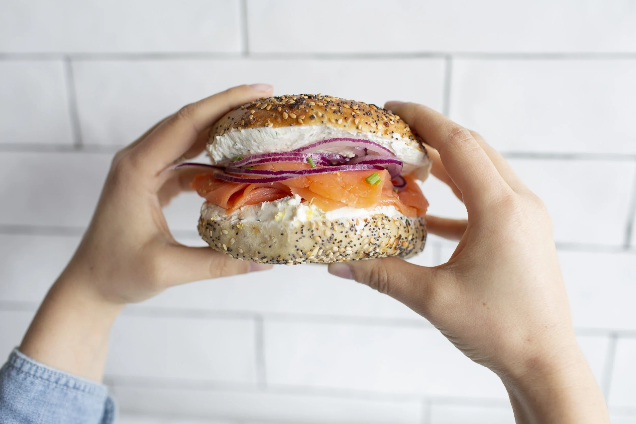Multi-seeded bagel loaded with lox, cream cheese, red onions and herbs, held by two hands, and ready to be eaten.