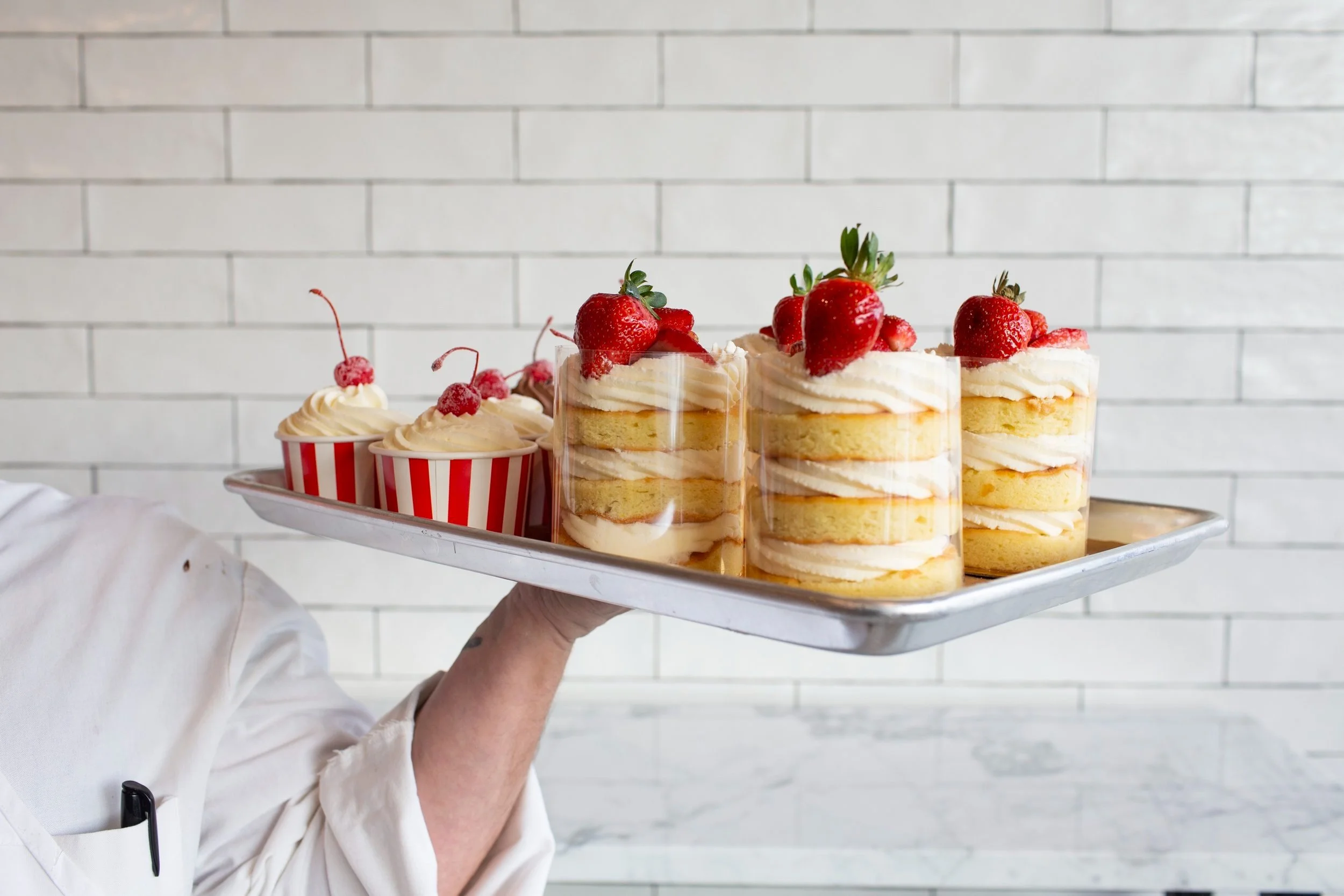 A bakery chef in a white coat holds a tray of red and white swirled cakes and treats featuring buttercream frosting, strawberries and cherries on top; the background is classic white NYC subway tile. 