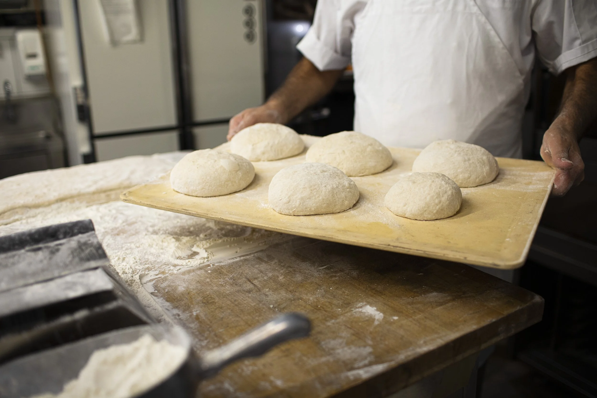 Wooden tray with pre-baked bread dusted with flour, being held by a chef in white jacket; a scoop of white flour in the foreground. 