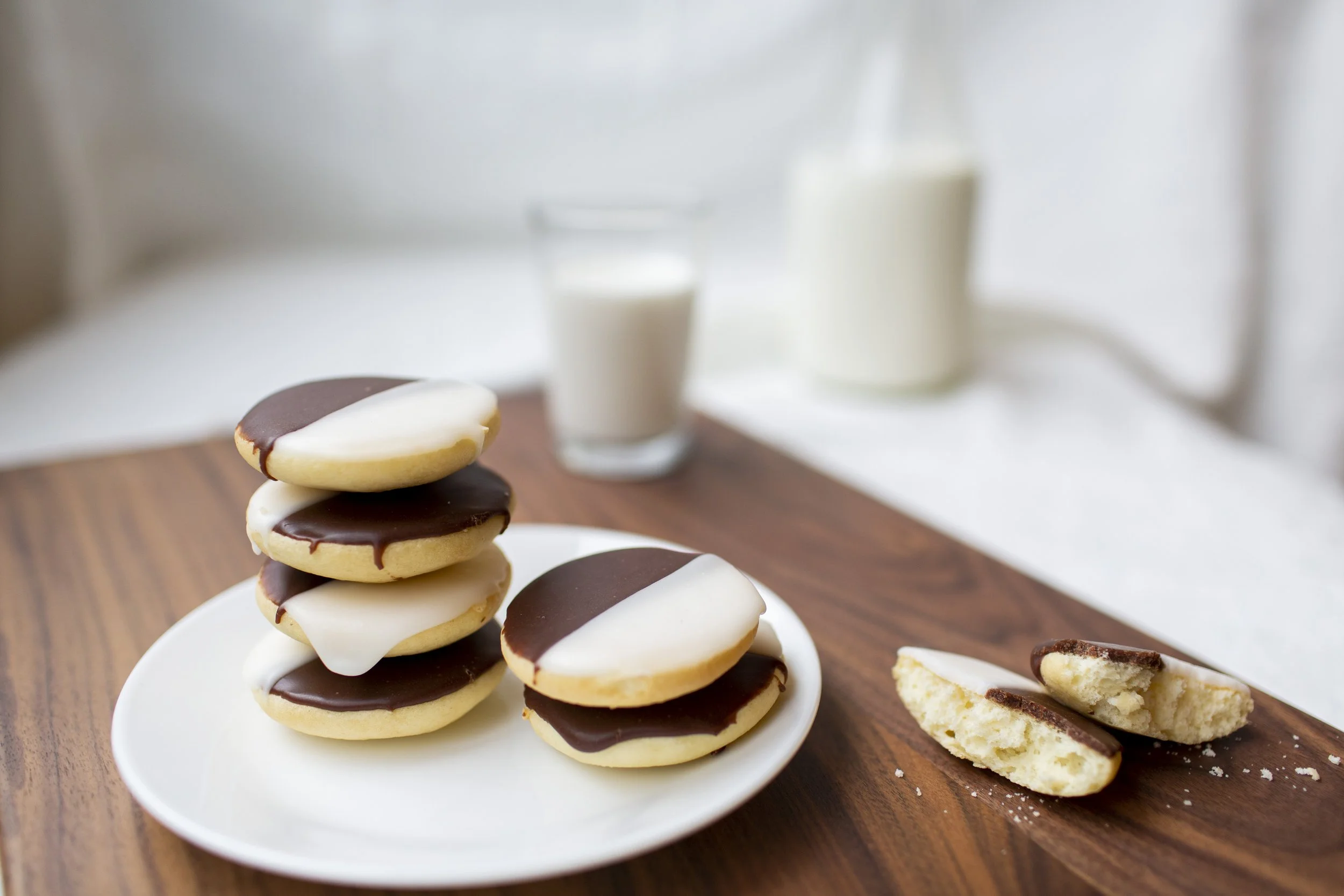 White plate with a stack of Black & White cookies sits on a wooden table in the foreground; glass of milk and glass jar of milk are in the background; Black & White cookie split in half with crumbs spill onto the table. 