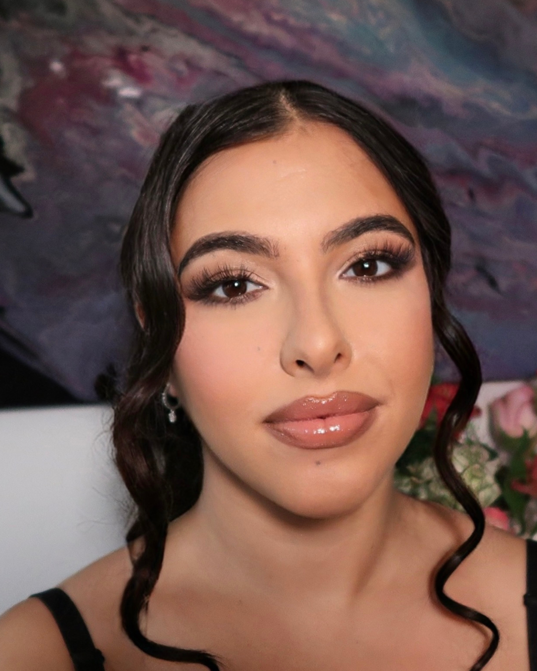 A young woman with long, wavy brown hair, wearing makeup with glossy lips, earrings, and a black top, posing in front of a colorful abstract painting and some pink flowers.
