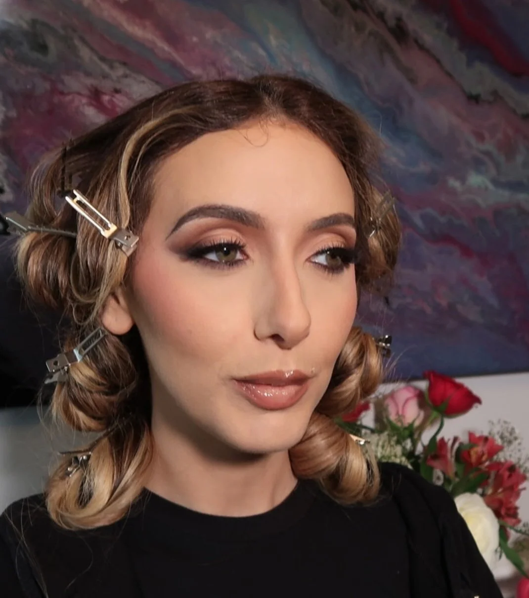 A woman with styled hair secured with hair clips, makeup, and a black top, is preparing for a photoshoot or event, with flowers in the background.