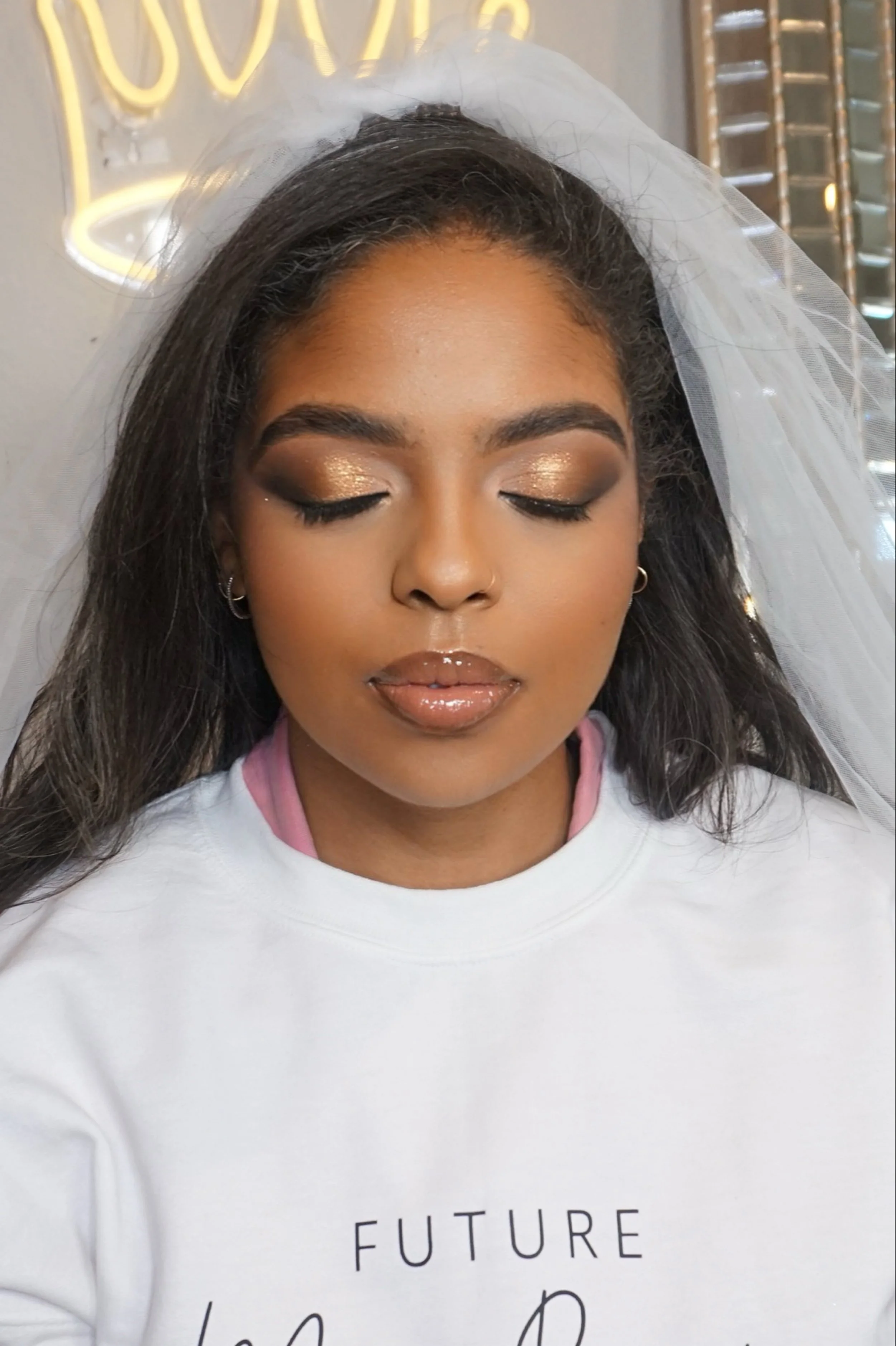 A woman with closed eyes, wearing makeup including glittery eyeshadow and glossy lips, with a white veil and a white shirt with black text that reads 'FUTURE'.