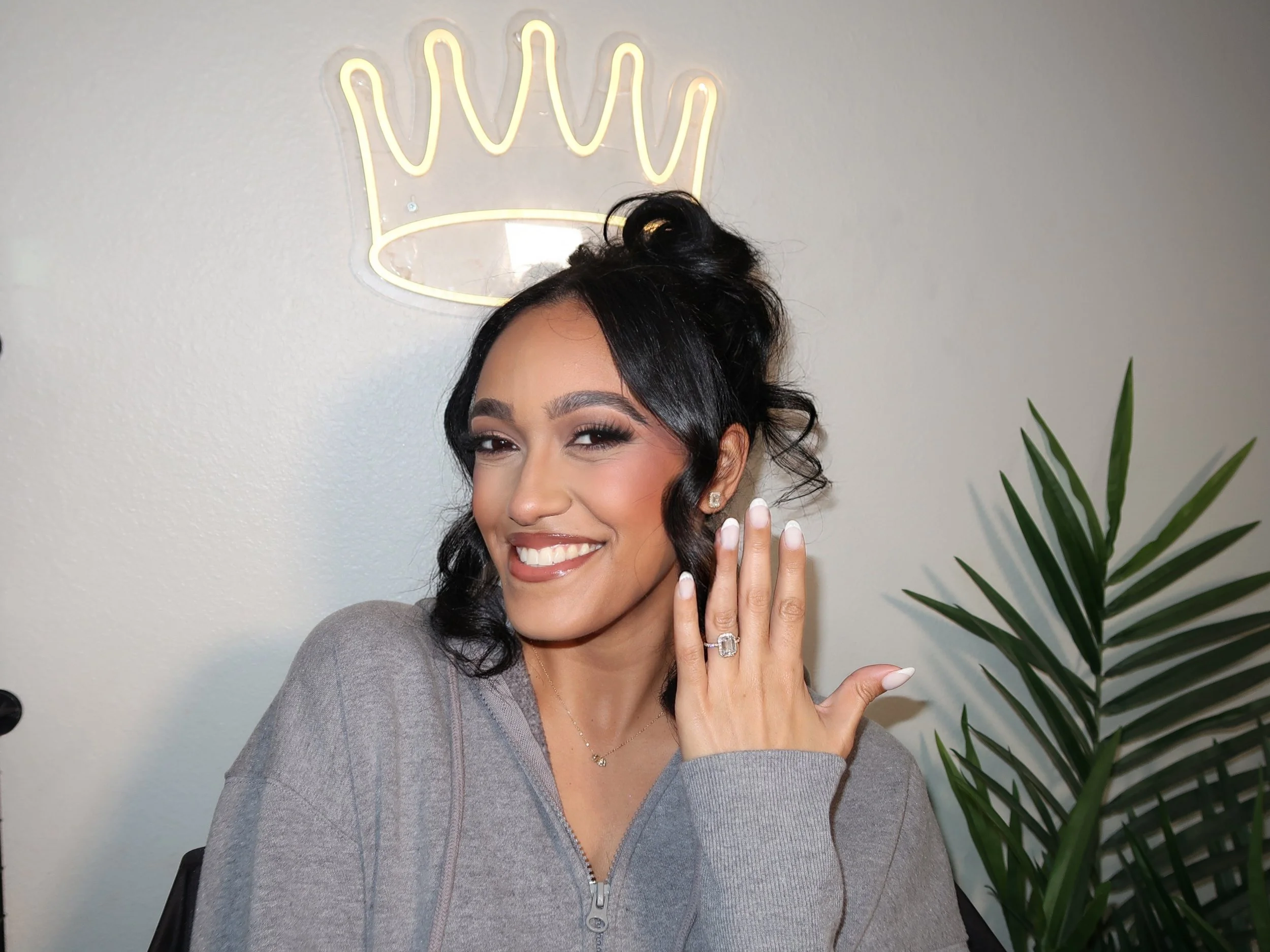 A woman with dark hair styled in loose curls, smiling and showing her perfect teeth, holding her hand near her face with a silver ring on her ring finger, standing in front of a wall with a neon crown sign above her head, a green plant is visible on the right side.
