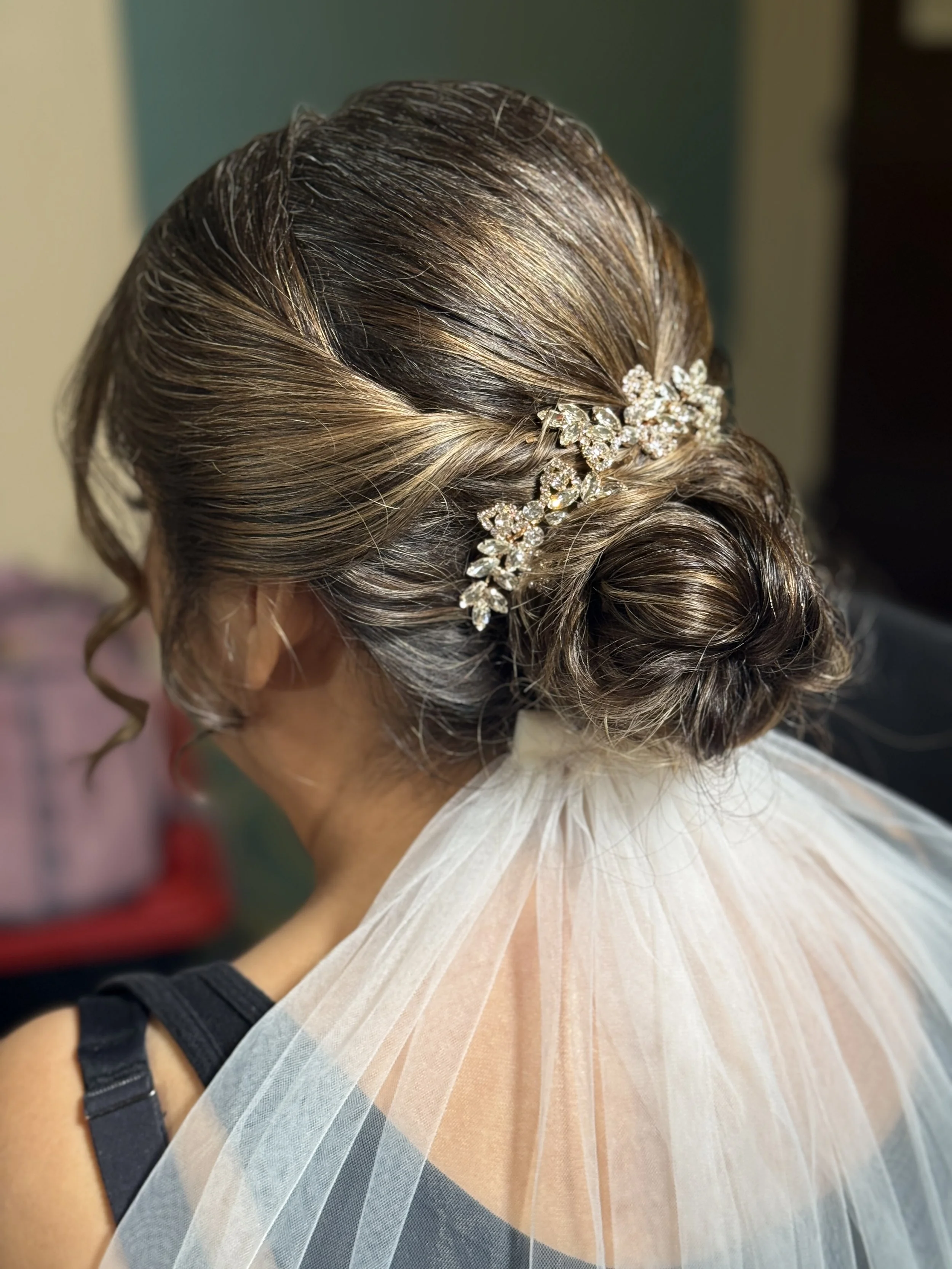 Close-up of an elegant updo hairstyle with a decorative floral hairpiece, featuring swirling curls and highlights, worn with a light-colored, textured fabric.