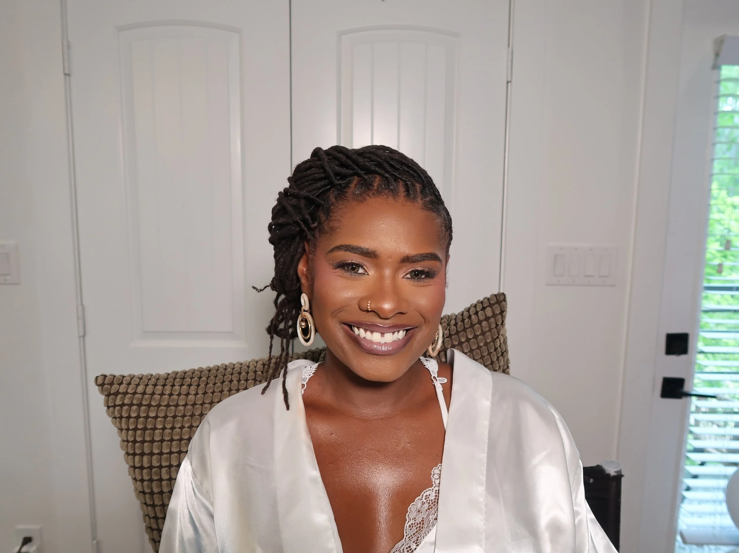 A woman with dark skin and braided hair styled in twists, smiling at the camera in a bright room with white walls, a window with greenery outside, and modern light switches.