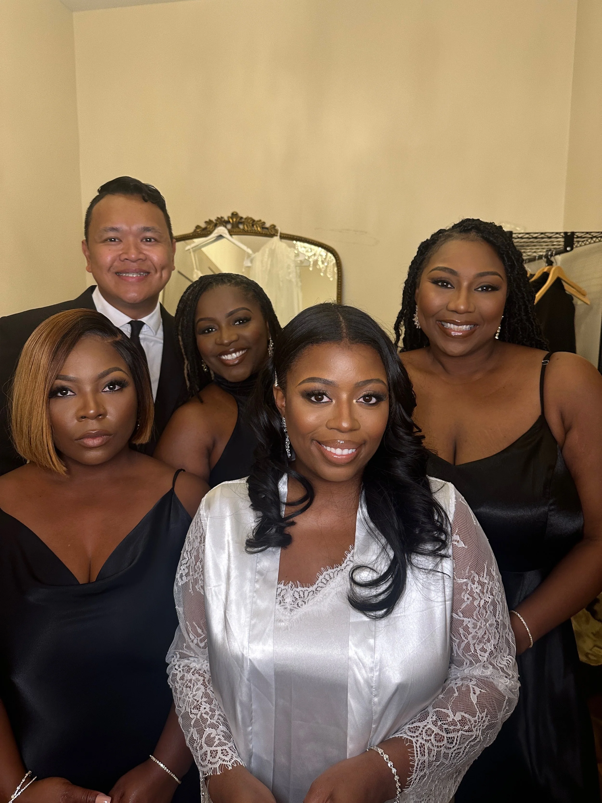 Group of six ethnically diverse people, five women and one man, dressed formally at an indoor event, with a mirror and clothing rack in the background.