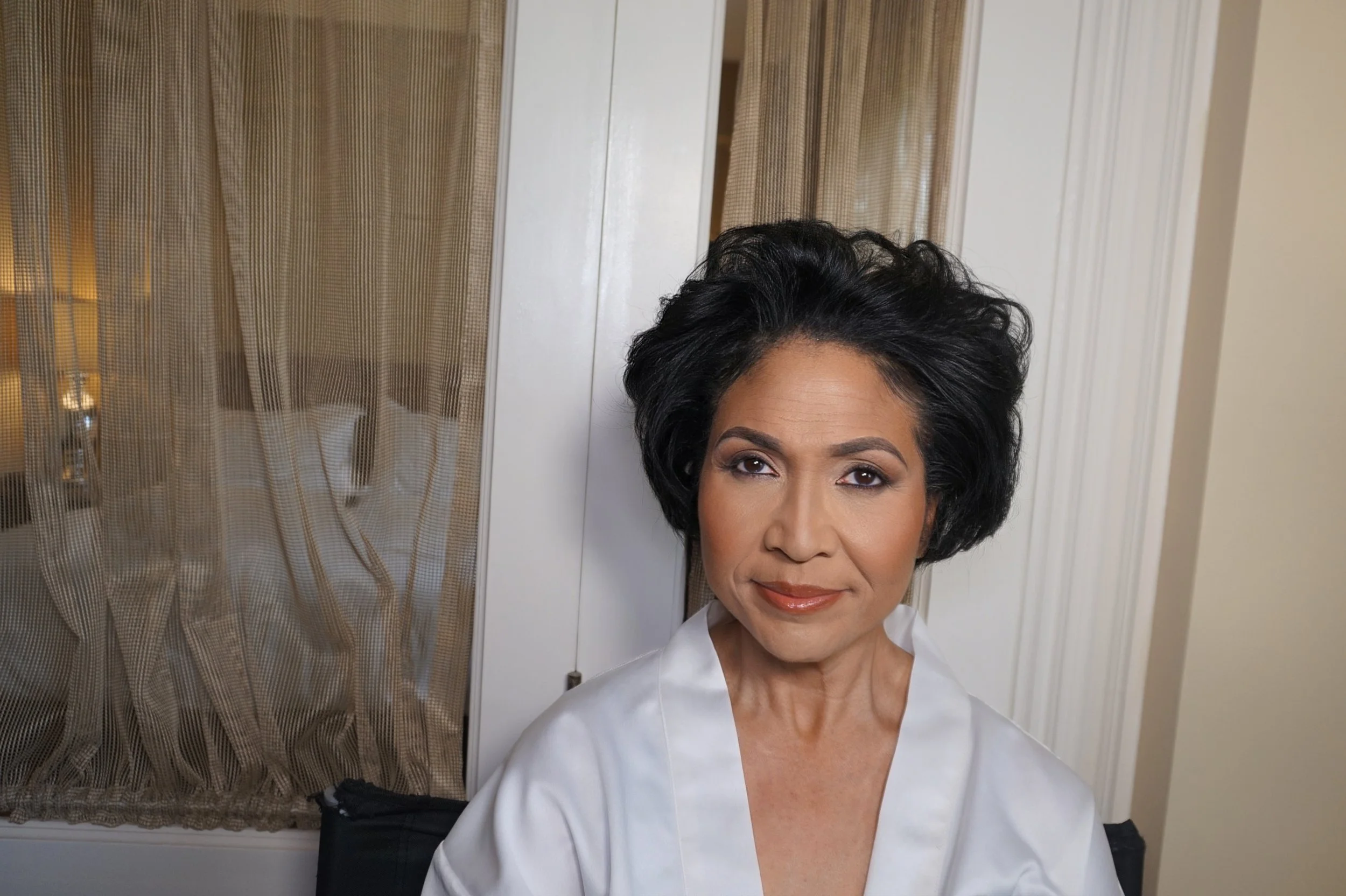 A woman with short black hair and makeup, wearing a white top, standing in front of a mirror with a closed door and beige curtains in the background.