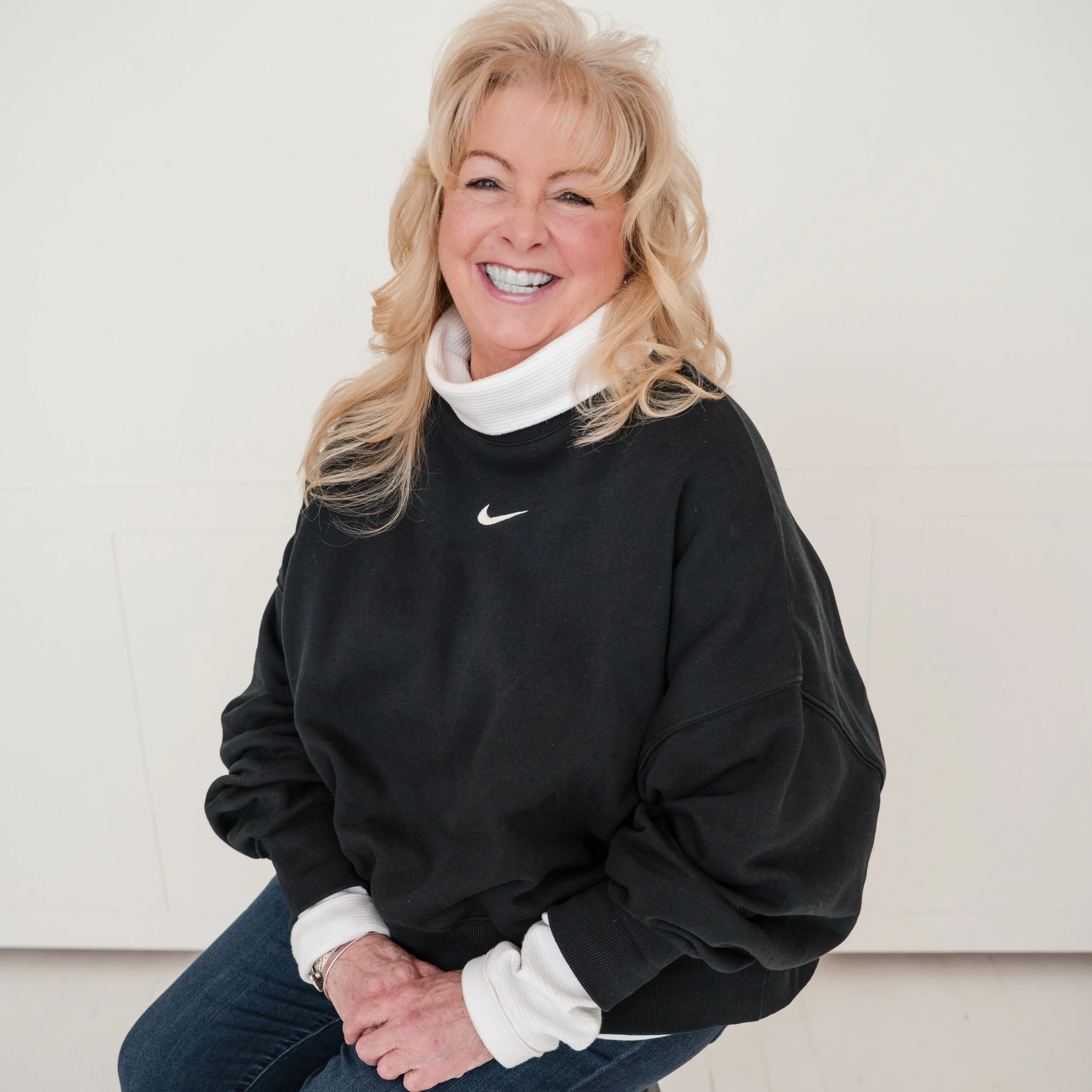 A smiling middle-aged woman with blonde hair sitting against a plain white background, wearing a black Nike sweatshirt, white turtleneck, and blue jeans.