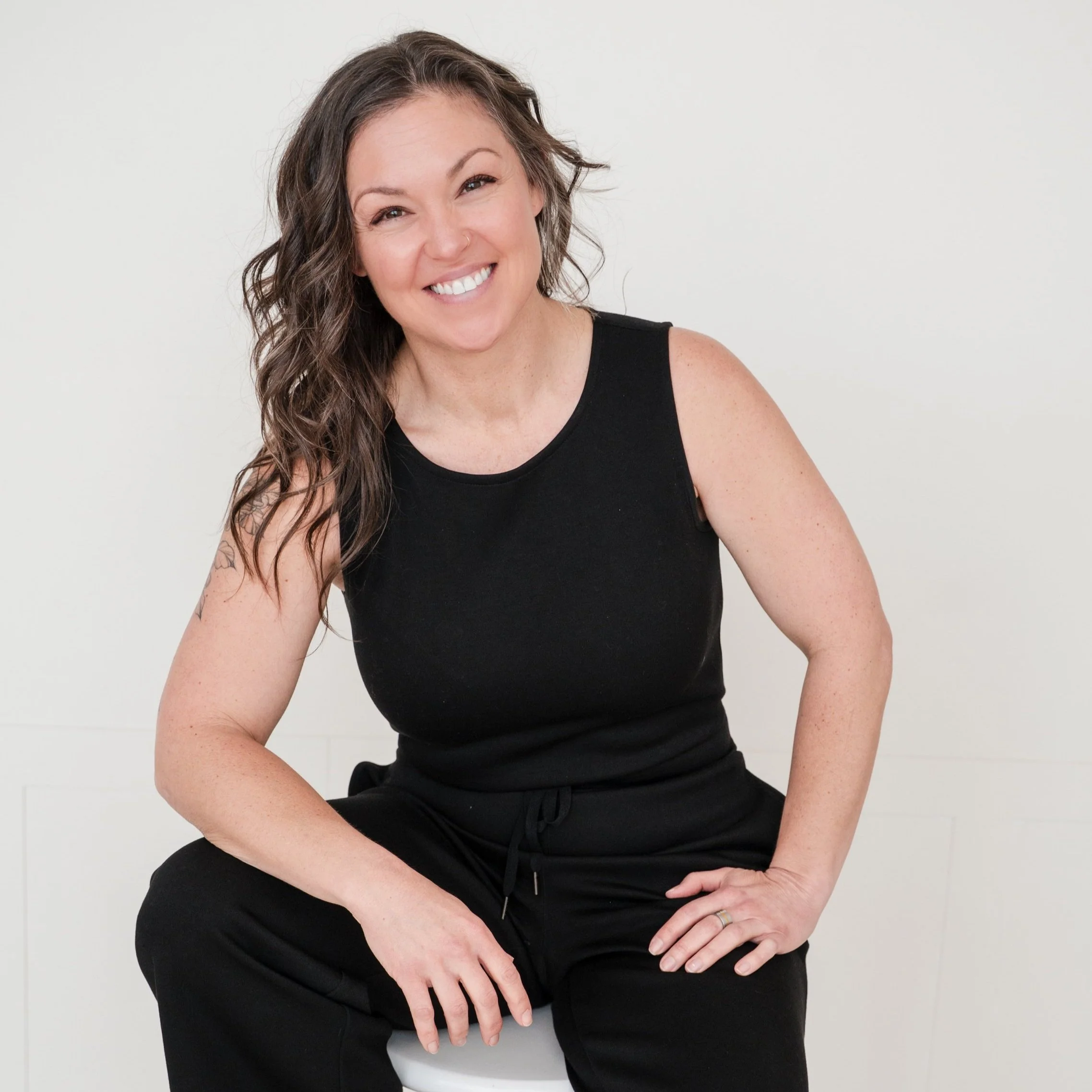 A woman with curly brown hair, smiling, wearing a black sleeveless top and black pants, sitting on a white stool against a plain white background.