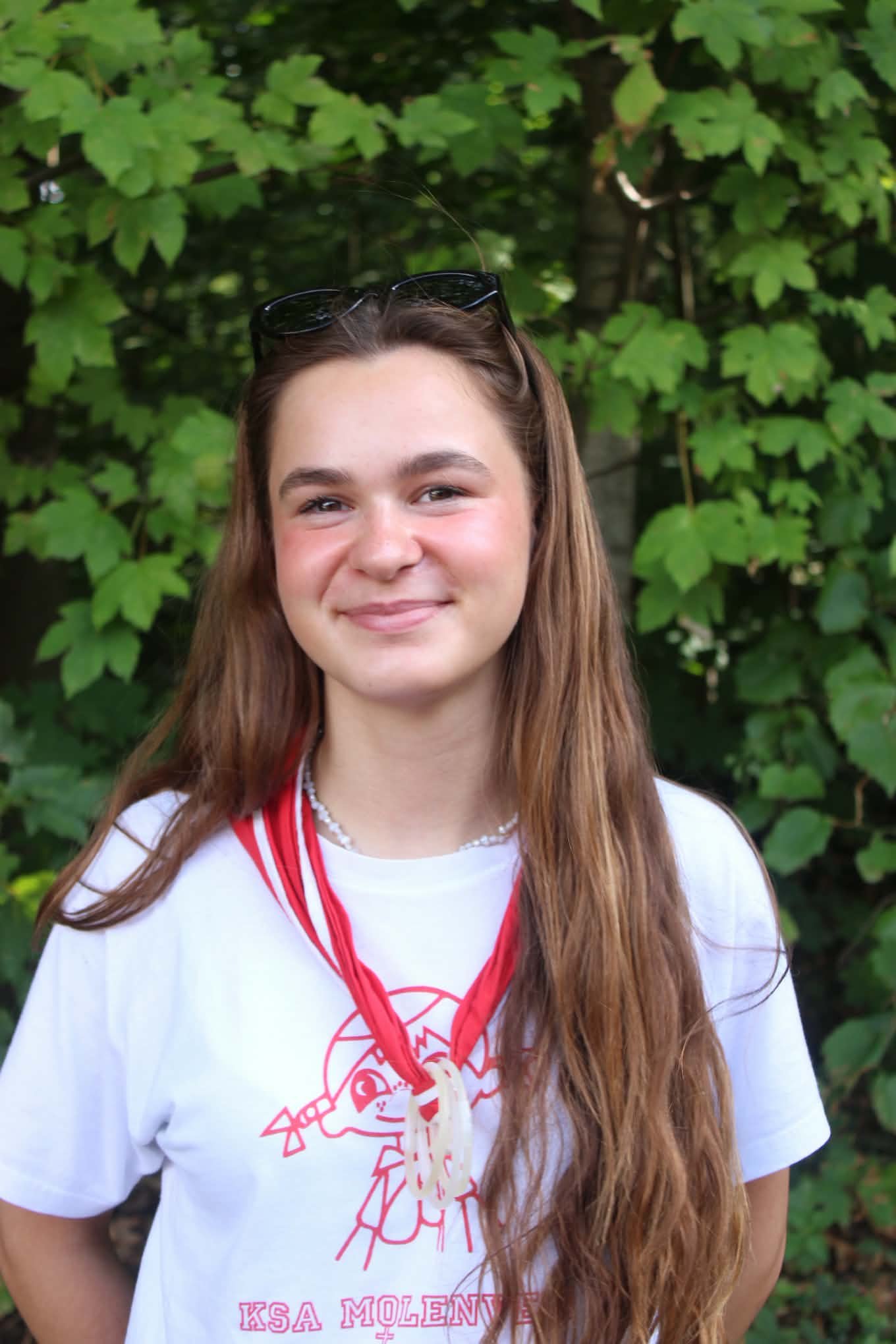 Young woman with long, wavy brown hair smiling at the camera, wearing a red hoodie with the name 'Louise' on it, outdoors with green foliage in the background.