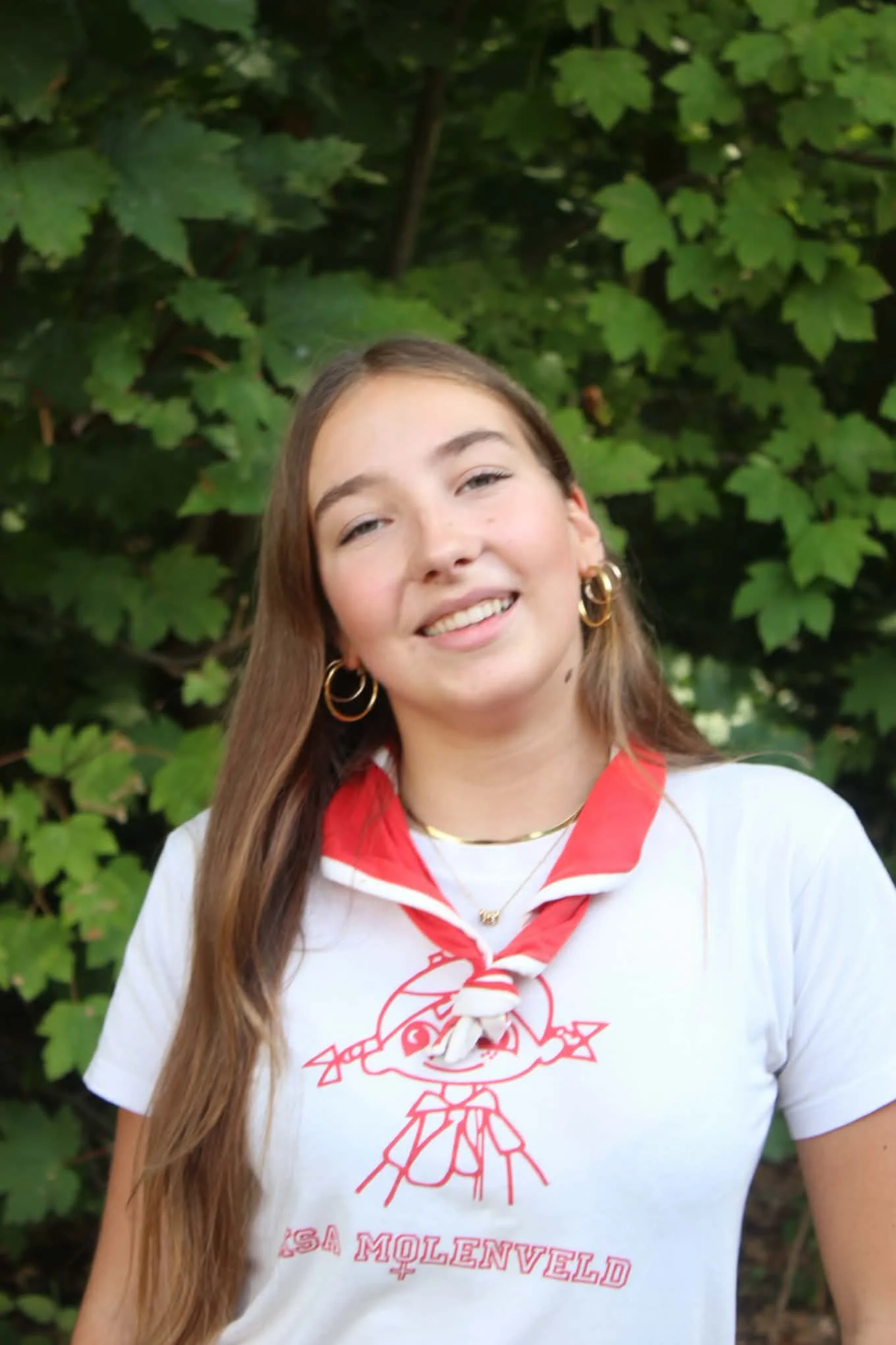 A young woman with blonde curly hair smiling outdoors against green leafy background, wearing a red hoodie with "SILKE" printed on it and a pink scarf around her neck.