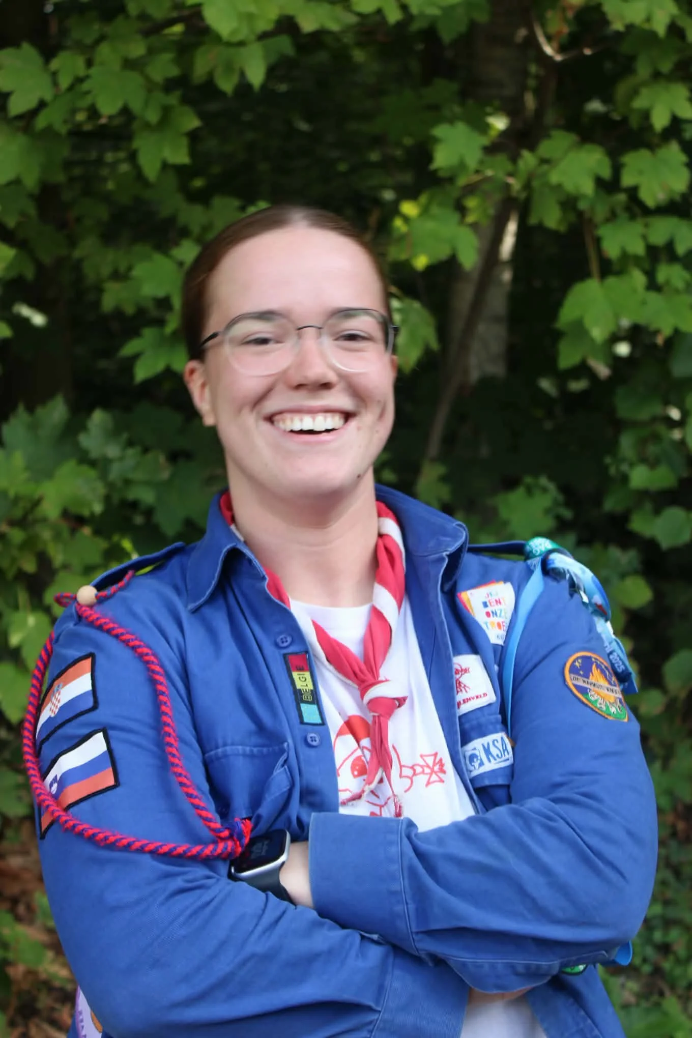A person with curly brown hair, glasses, and a nose piercing, wearing a red hoodie with "ORIANE" written on it and a red and white neckerchief, standing outdoors in front of green leafy foliage.