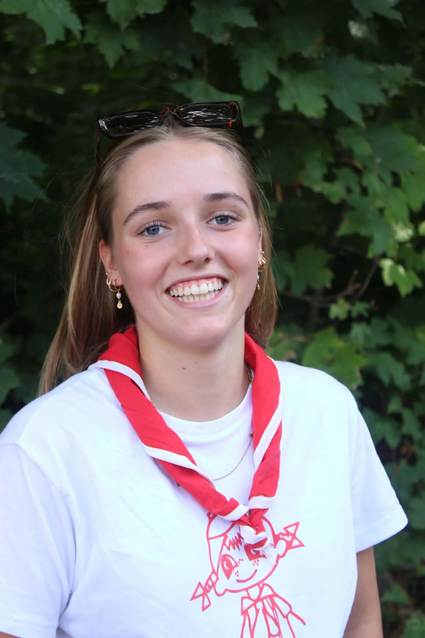 A young woman with long, wavy light brown hair and blue eyes smiling, wearing a red hoodie, standing outdoors in front of green leafy bushes.