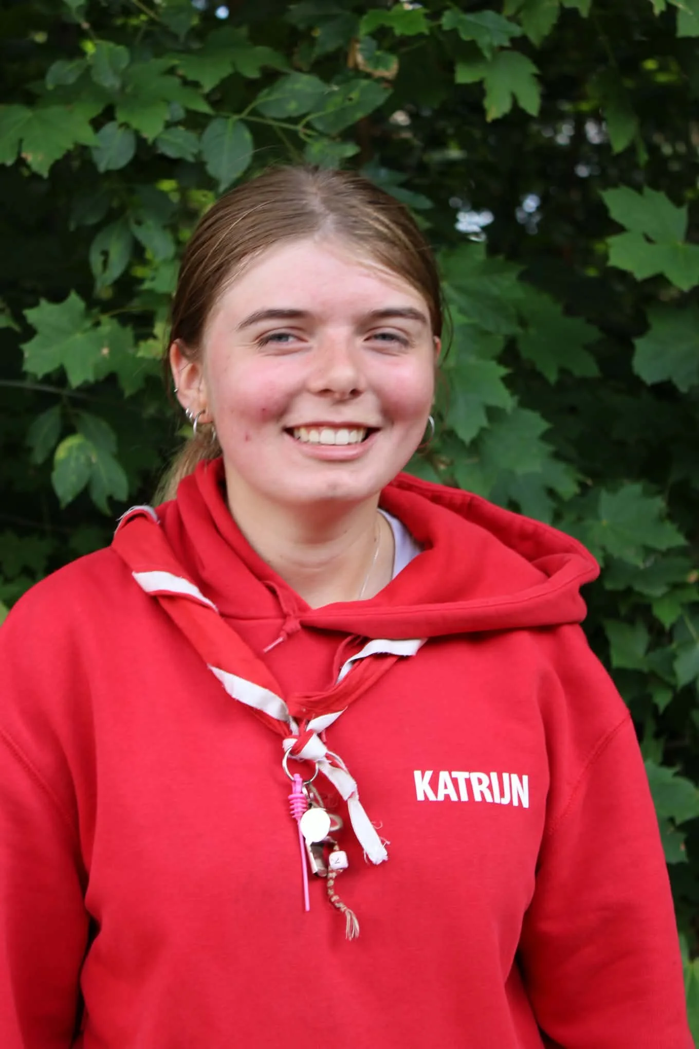 A smiling person with glasses, wearing a red hoodie and a blue jacket, stands in front of green foliage.