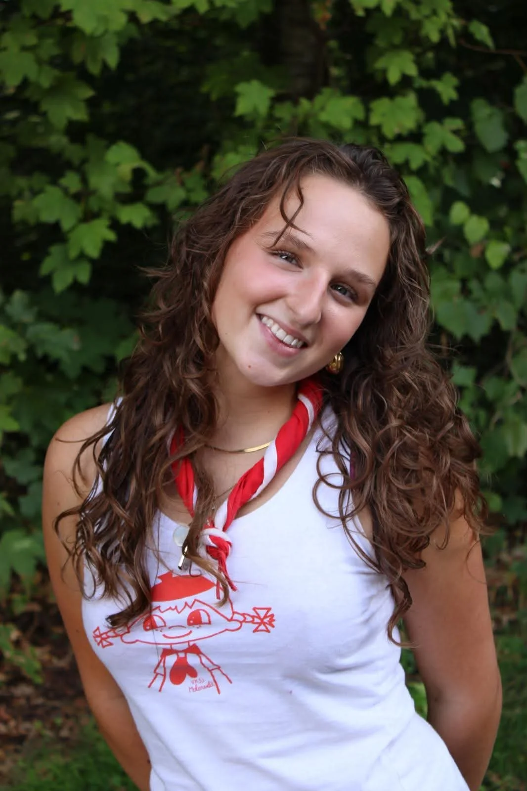 A young girl in a red hoodie and blue scout jacket standing outdoors in a wooded area, smiling with arms outstretched.