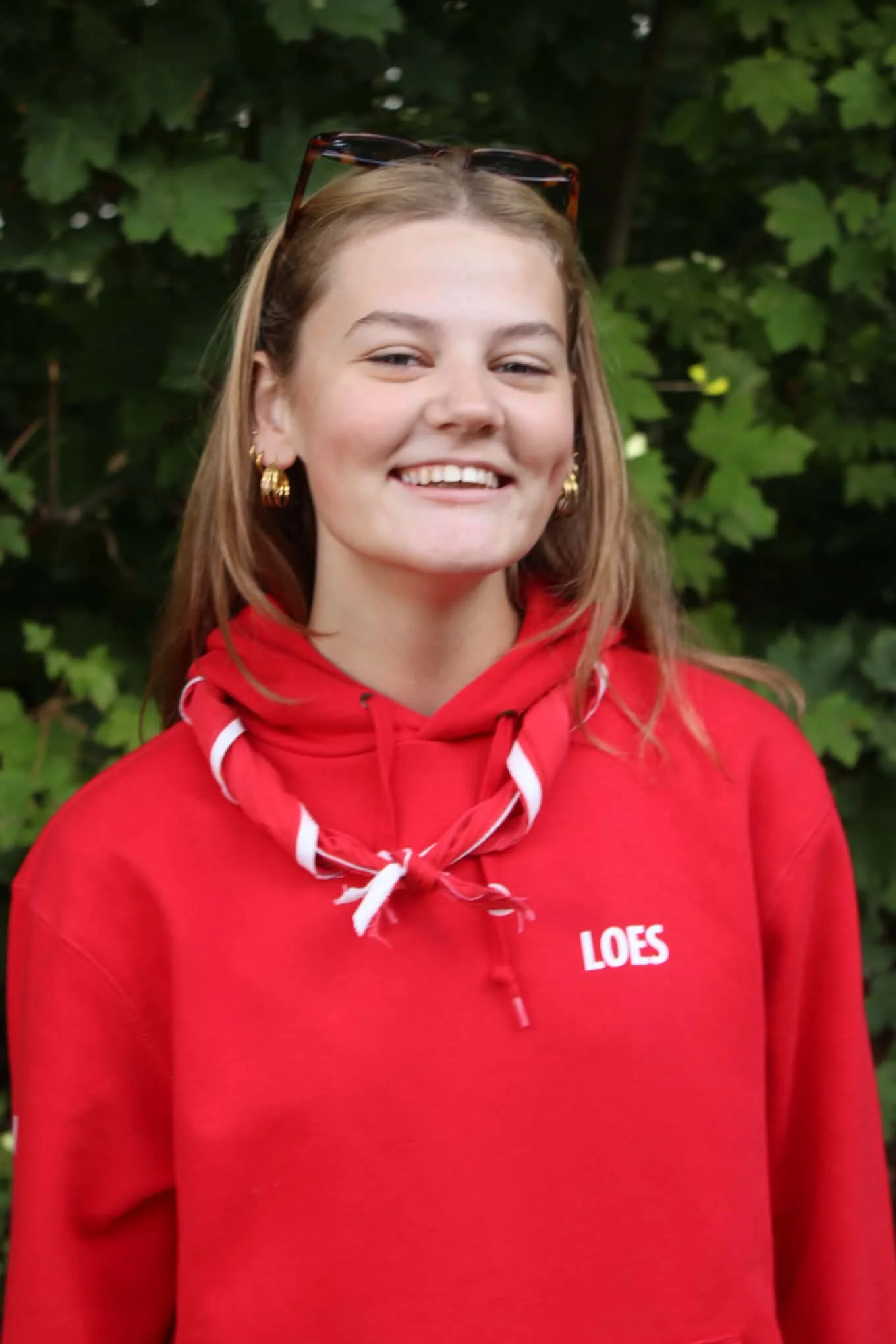 Young woman with long reddish hair smiling, wearing a red hoodie with white lettering, standing in front of green leafy bushes.