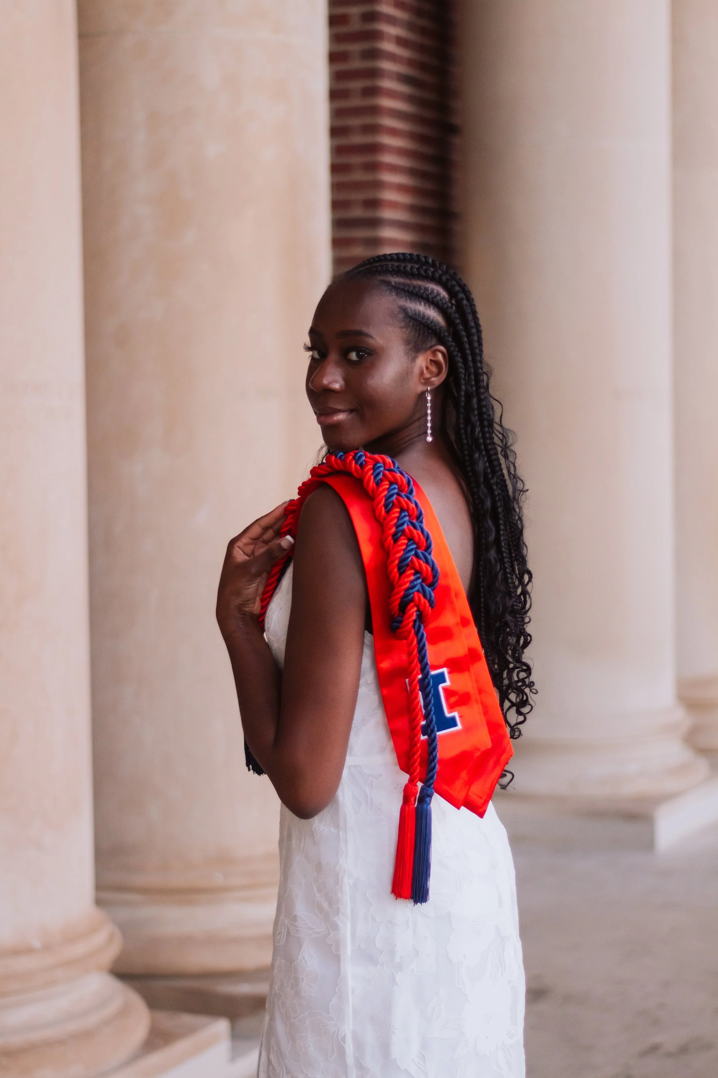 Young woman in a white dress with braided hair, wearing a red and blue sash, standing outdoors with columns and brick pillars in the background.