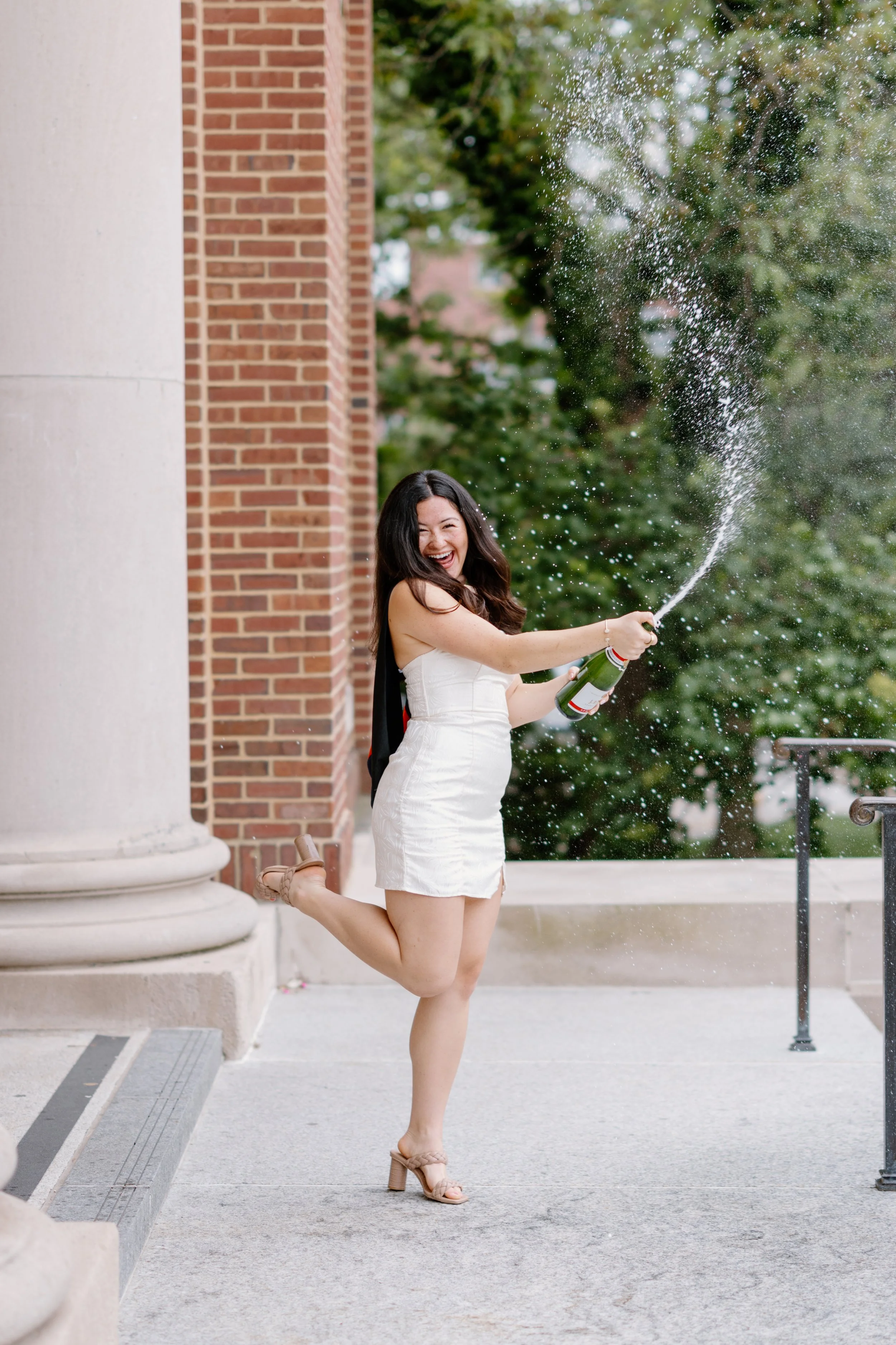 A woman in a white dress and high heels is celebrating outdoors, spraying champagne and kicking her leg up with a big smile.