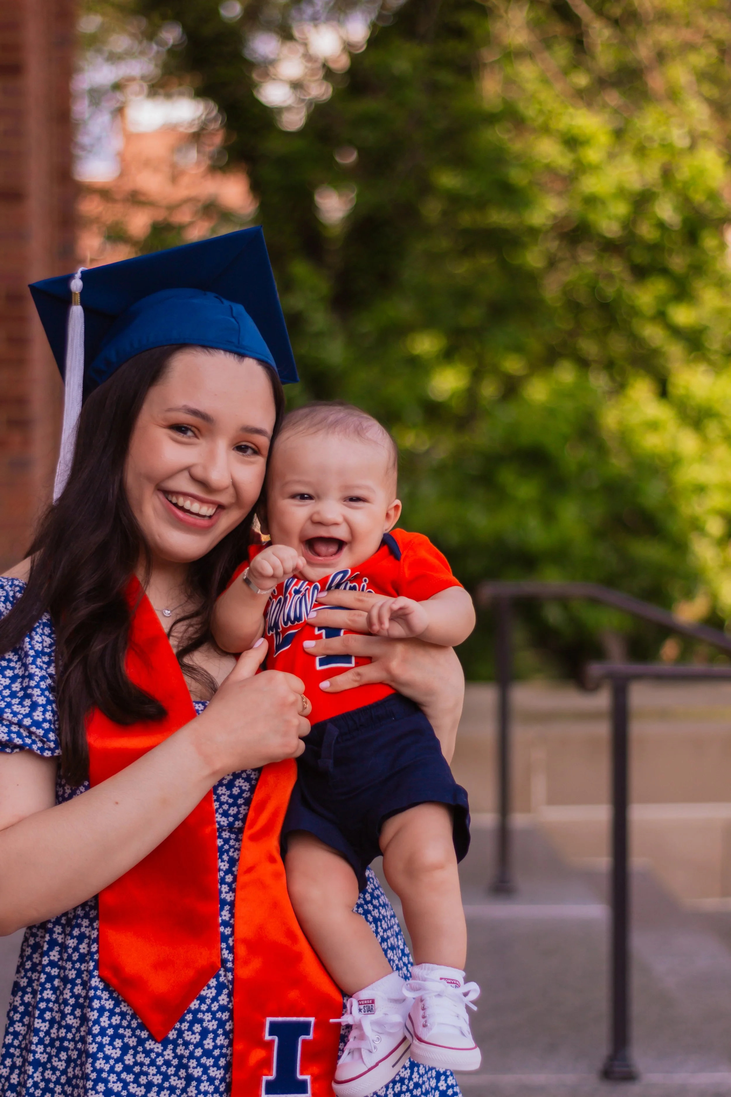 A young woman in a blue graduation cap and orange stole holding a smiling baby boy dressed in red and navy blue outdoors with trees in the background.