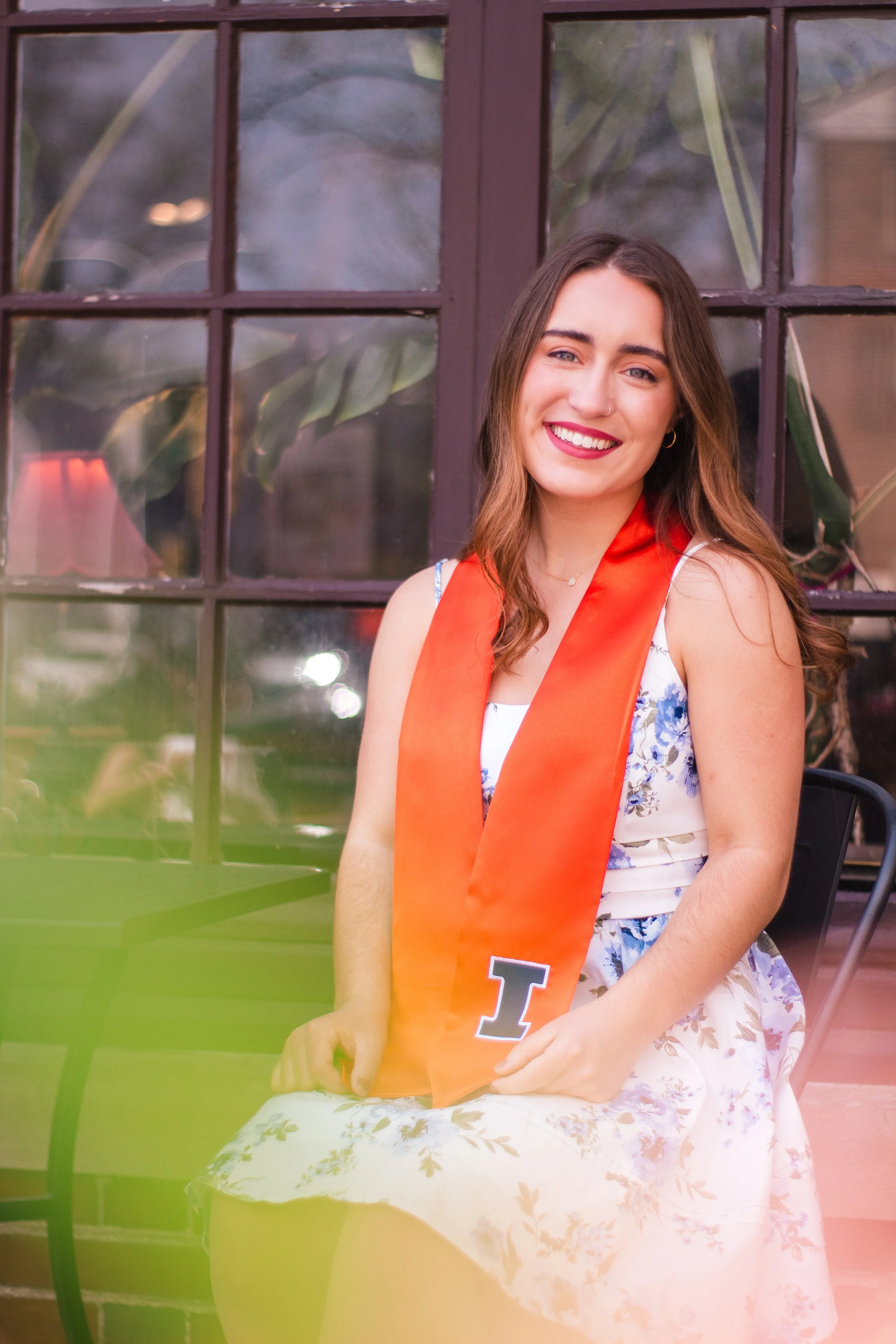 A young woman with long brown hair, wearing a floral dress and orange graduation stole with the letter 'I', sitting outside near a glass door, smiling at the camera.