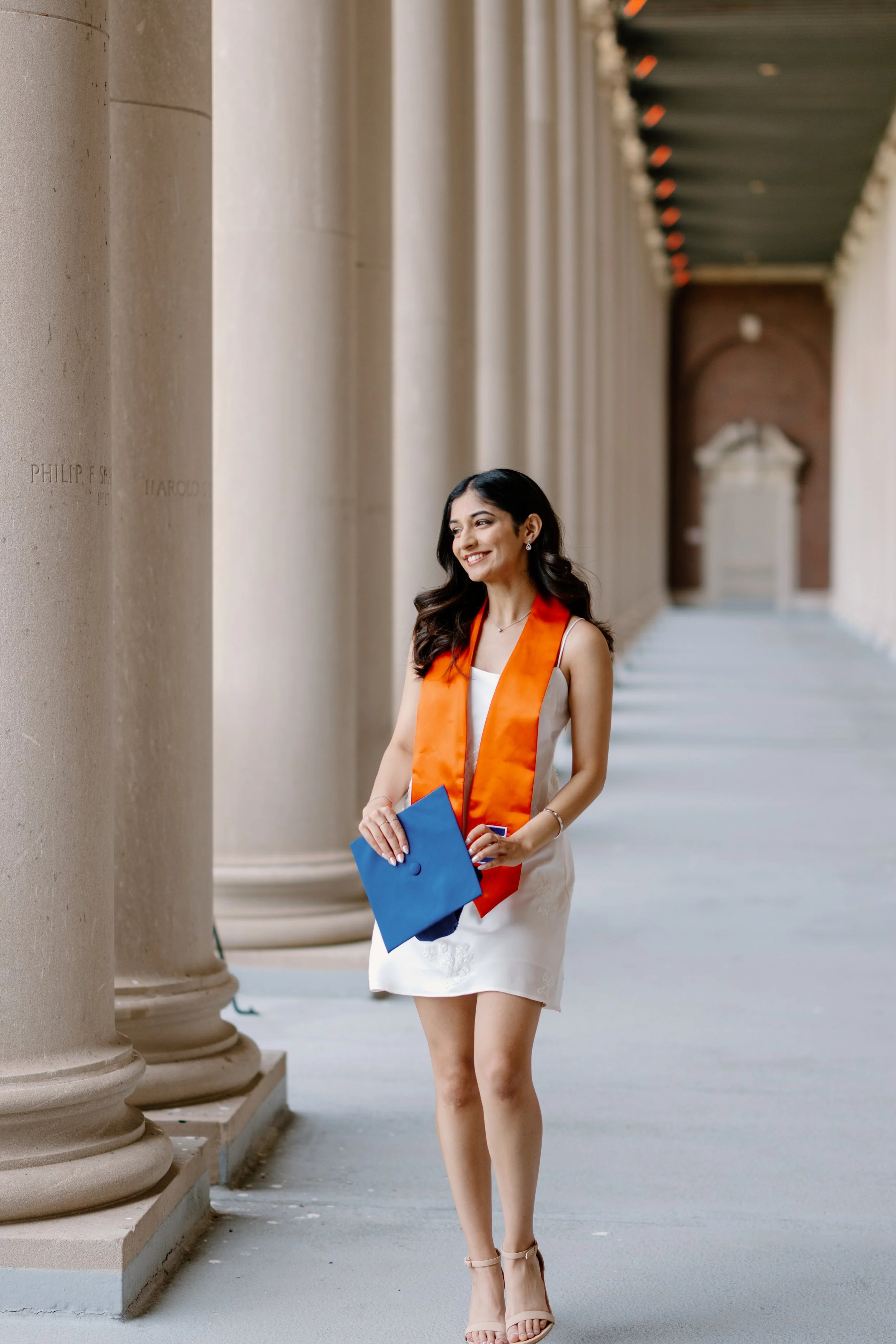 Young woman in white dress with an orange graduation stole, holding a blue diploma cover, smiling, walking in a colonnade.