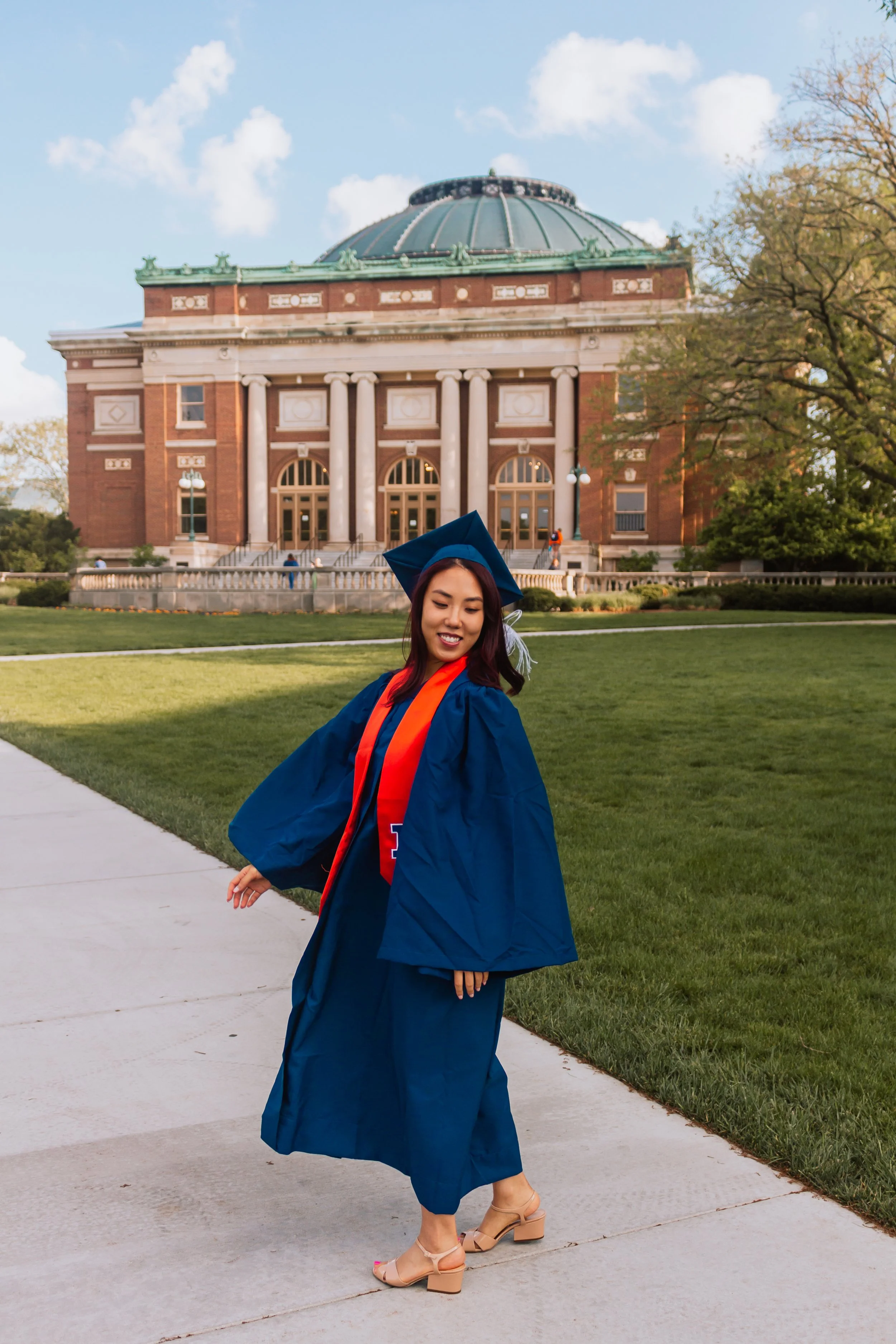 A young woman in graduation cap and gown walking on campus near a large, historic building with columns and a domed roof, smiling and looking down.
