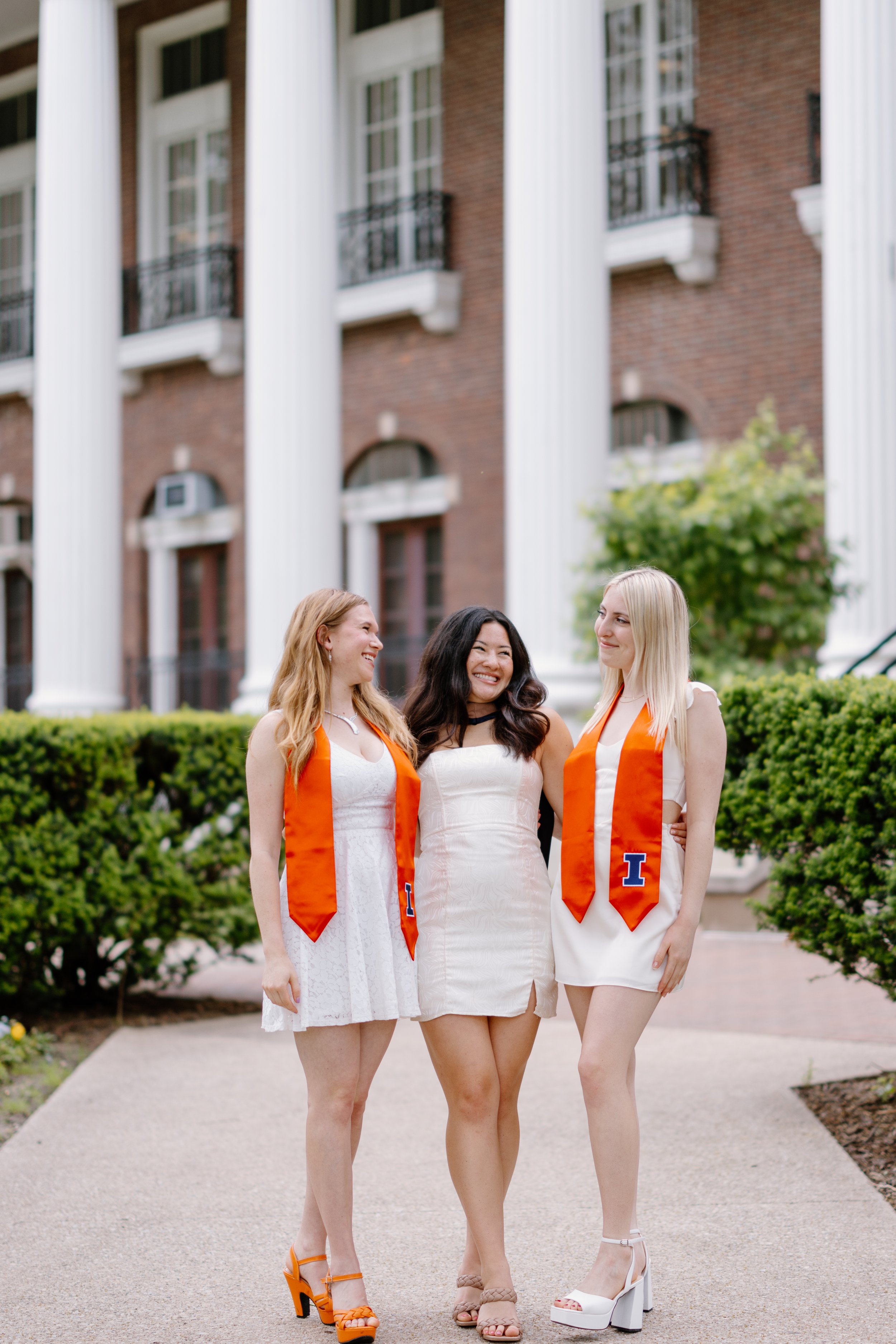 Three young women in white dresses with orange graduation stoles standing outside in front of a brick building with white columns, smiling and looking at each other.