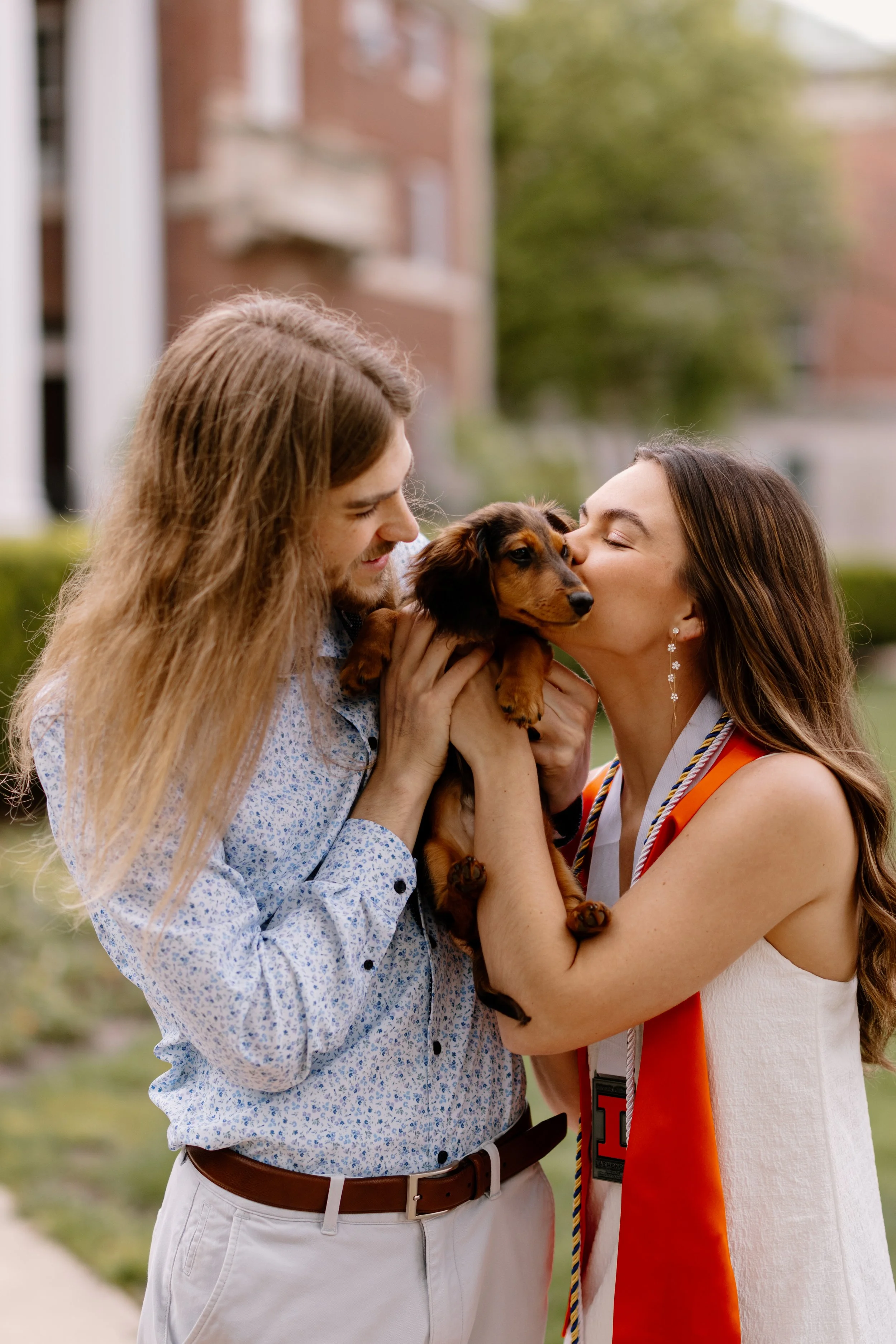 A woman in a white dress and a woman in a graduation gown kissing a small brown and black dachshund puppy with a man with long hair holding the puppy and smiling.