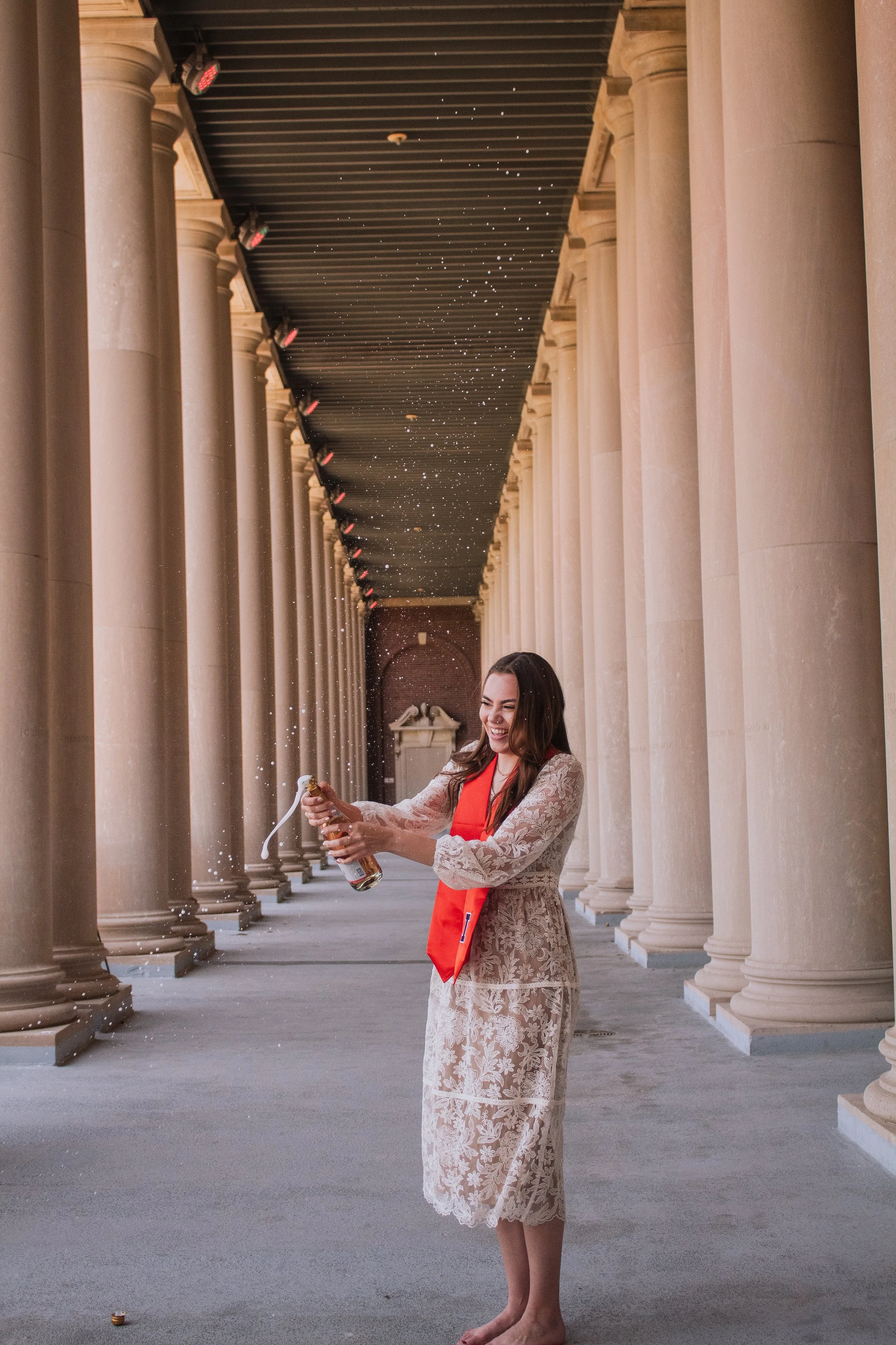 Woman in lace dress with red sash celebrating, opening a champagne bottle in front of large classical columns, with champagne spray in the air.