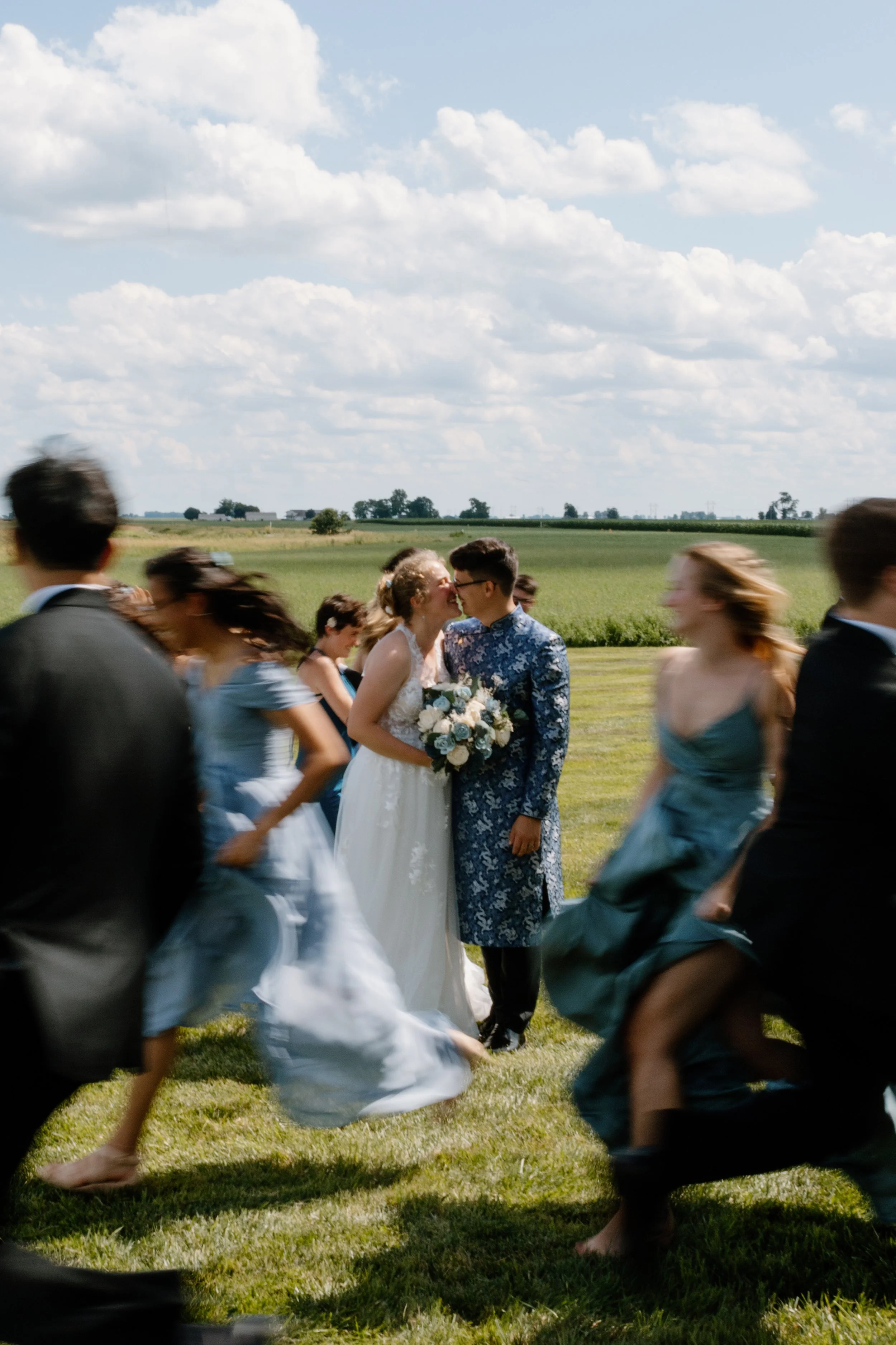A bride and groom kissing in the center of a group of wedding guests outdoors on a sunny day with green fields and a blue sky in the background.