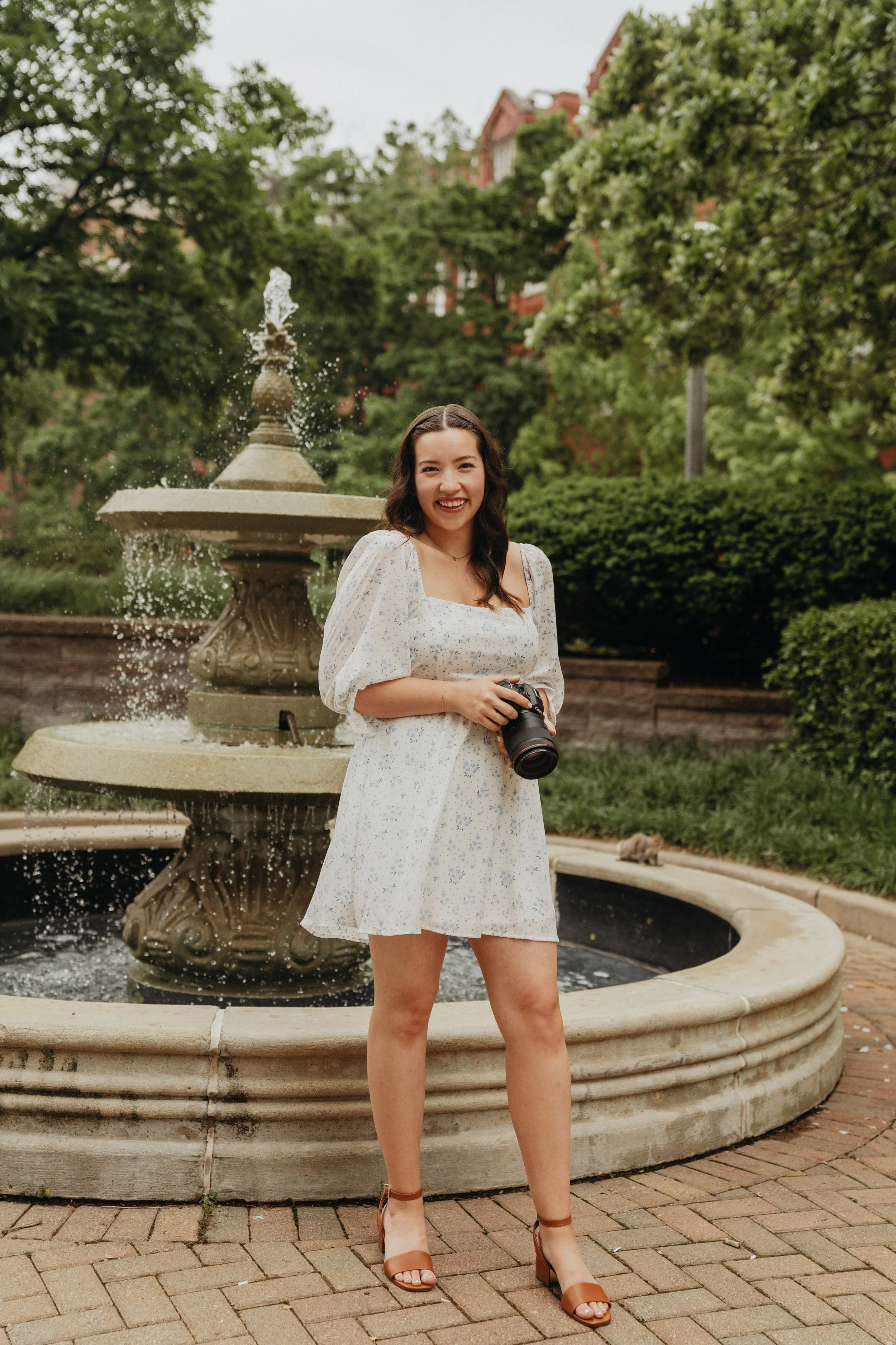 A young woman in a white floral dress and brown heels standing in front of a fountain in a park, holding a camera and smiling.