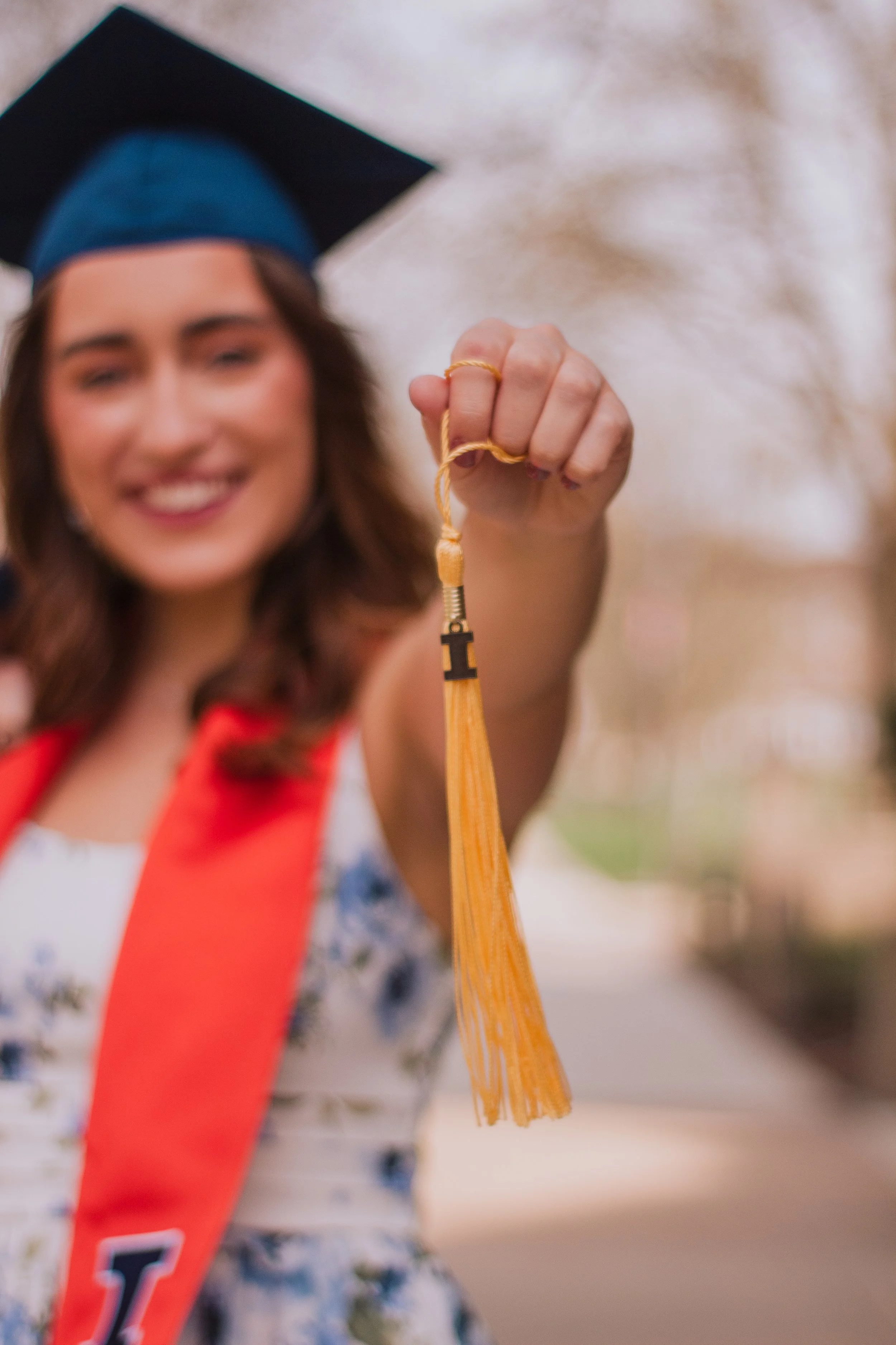 Smiling woman in graduation cap holding a gold tassel in focus, blurred background.