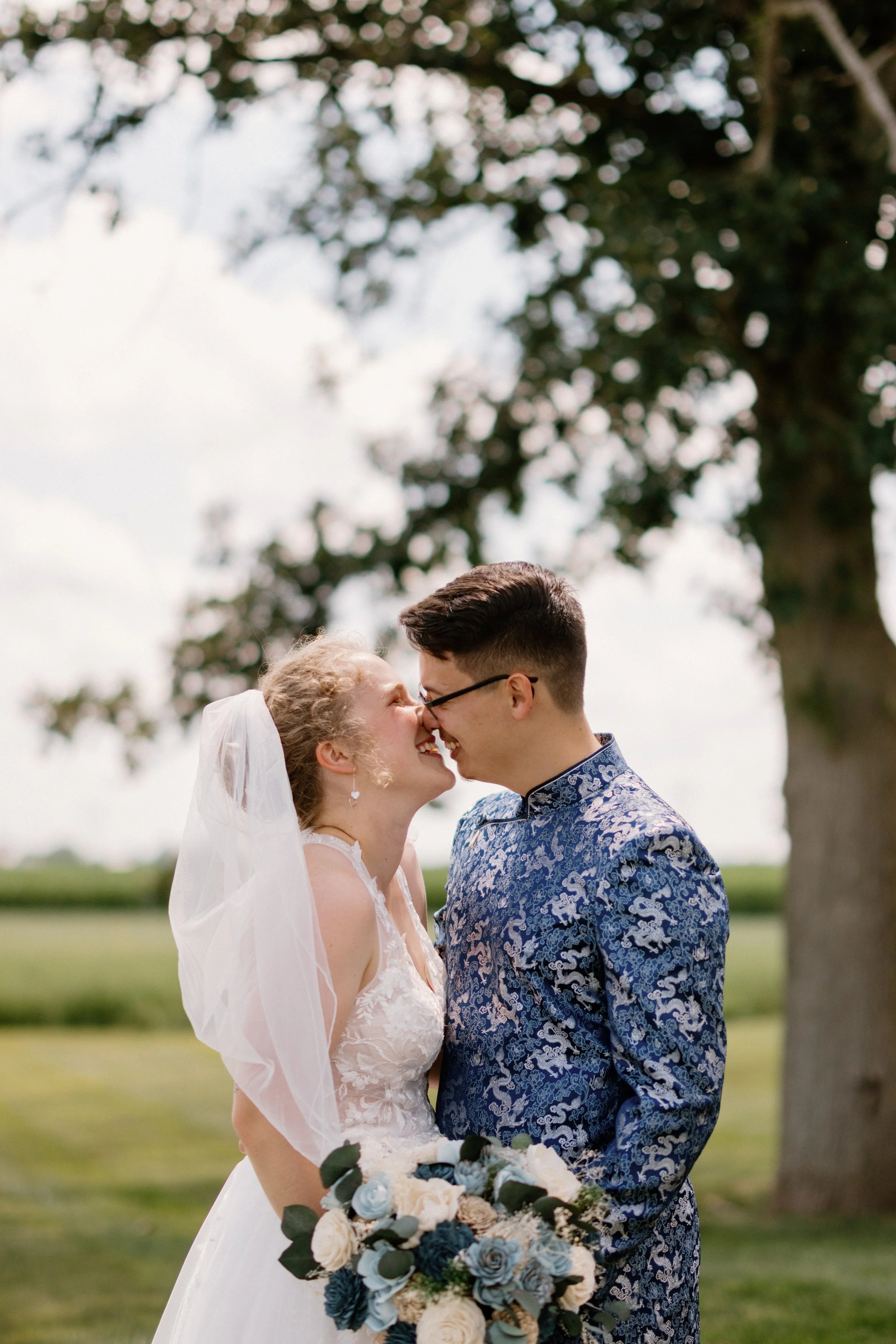 A bride and groom smiling and touching noses outdoors on their wedding day, holding a bouquet of white and blue flowers with greenery, with trees and a partly cloudy sky in the background.
