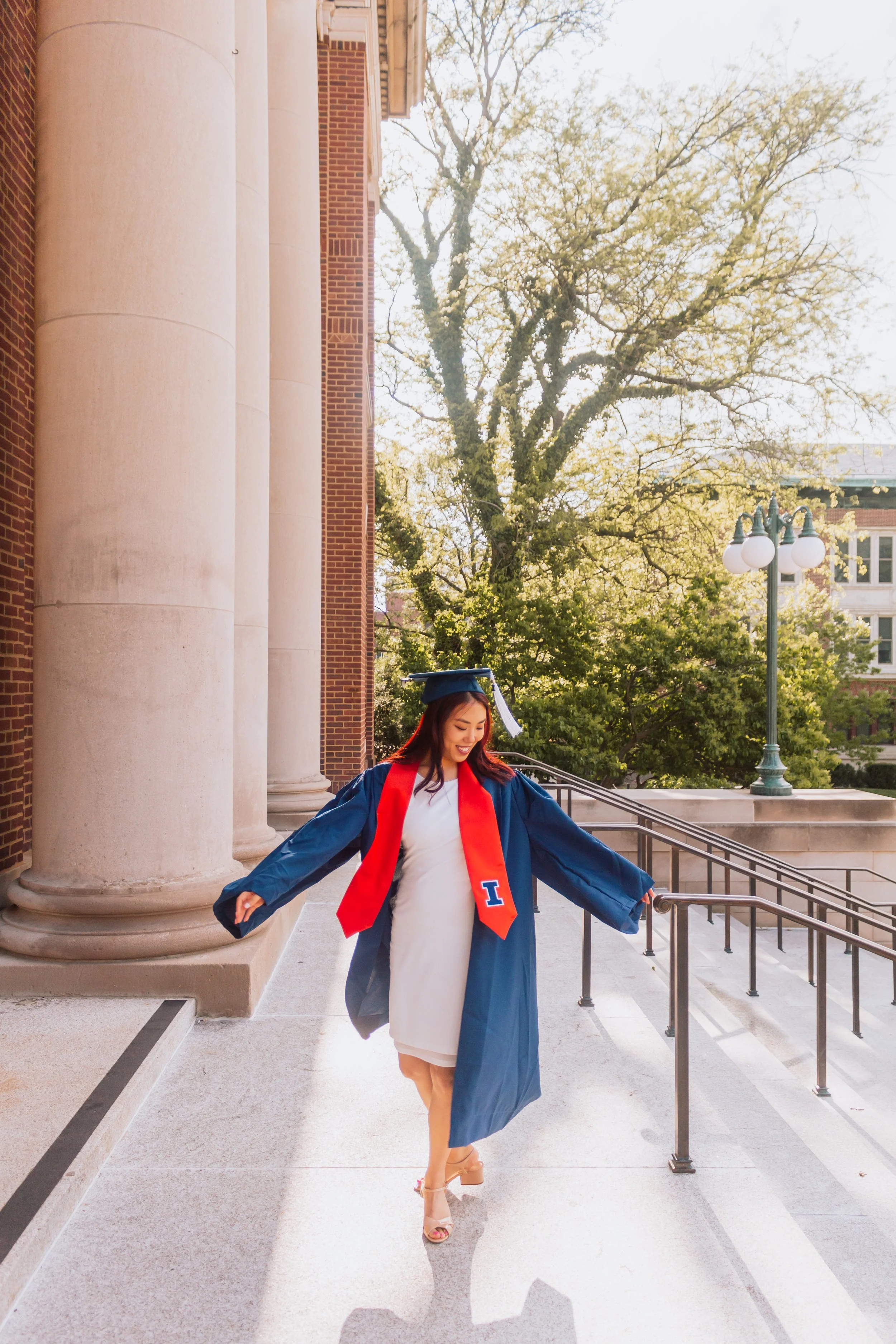 A woman in a graduation cap and gown with a red stole walks down the steps outside a building with large columns and trees in the background.