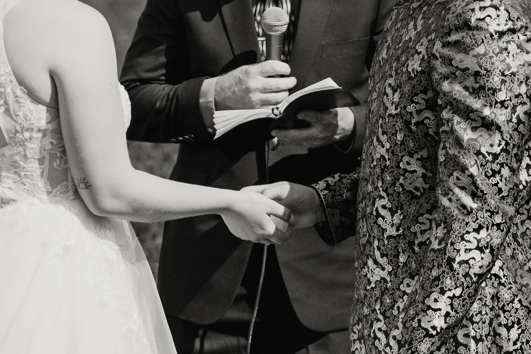 A couple holding hands during a wedding ceremony, with officiant reading from a book in the background.