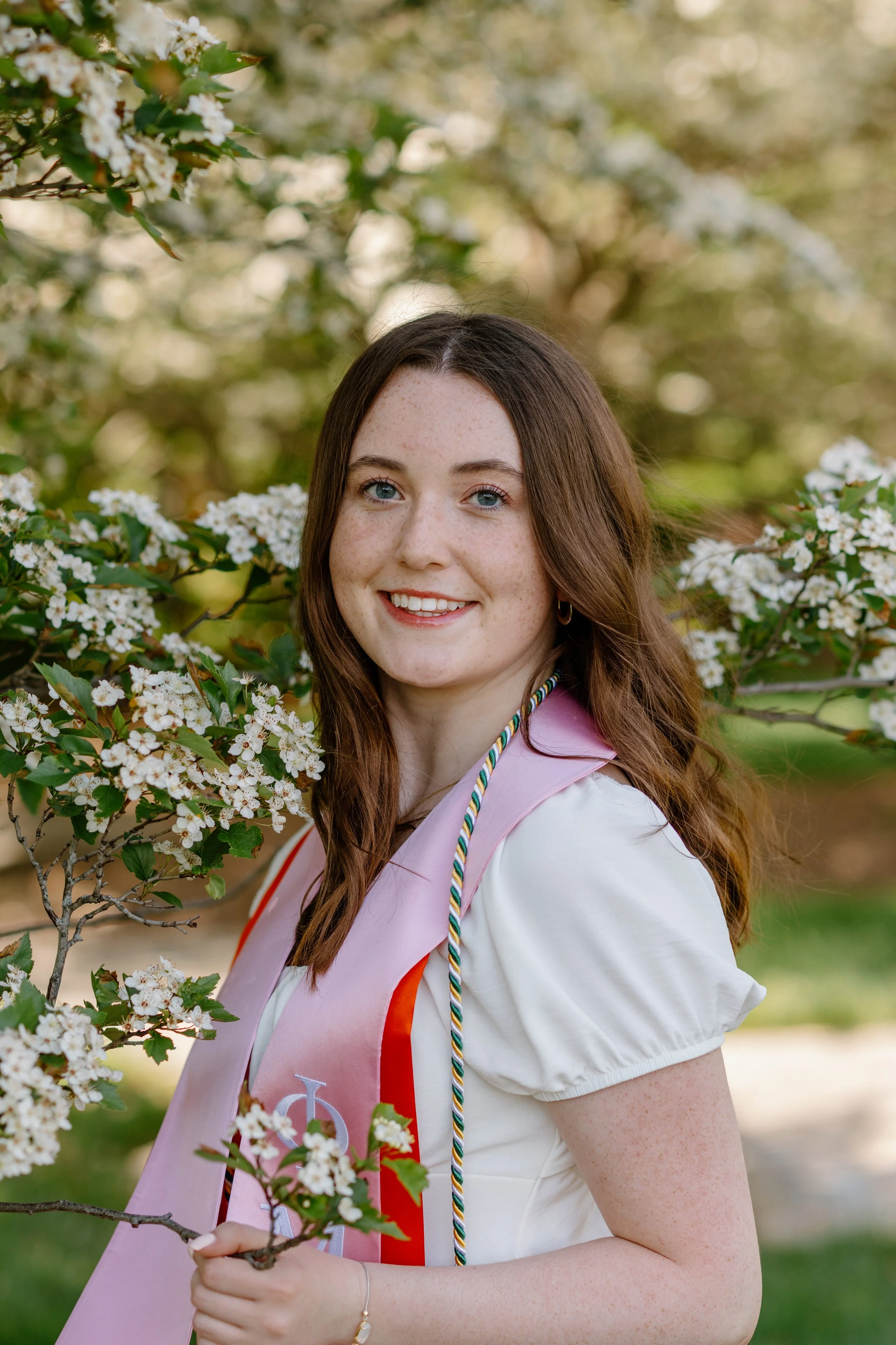 A young woman with long brown hair, blue eyes, and freckles, smiling outdoors, holding a branch with white blossoms, wearing a pink and red graduation stole and a white dress.