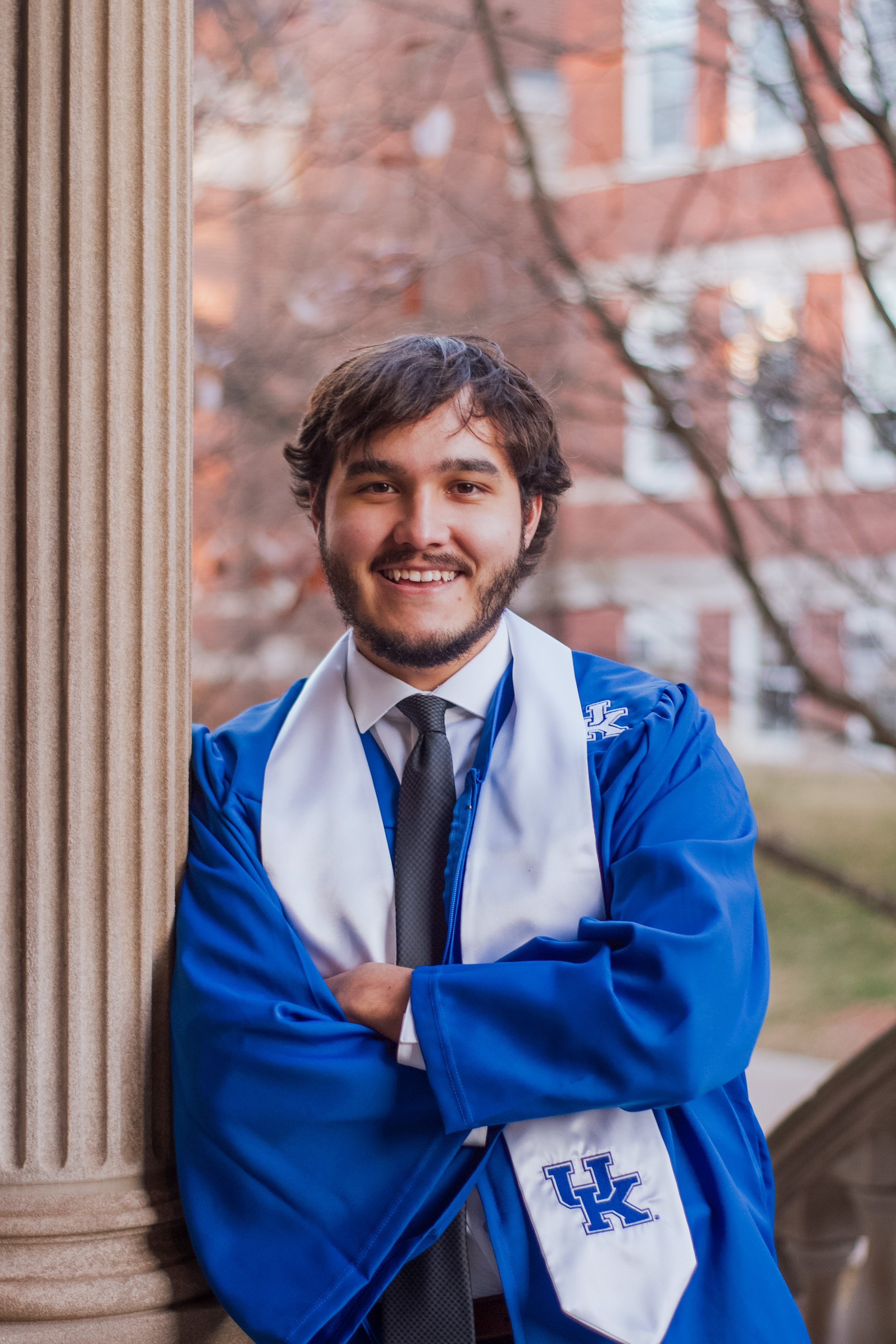 A young man with brown hair and a beard in a blue graduation gown and white stole stands outdoors, smiling and leaning against a stone column. A building with red brick walls and trees can be seen in the background.