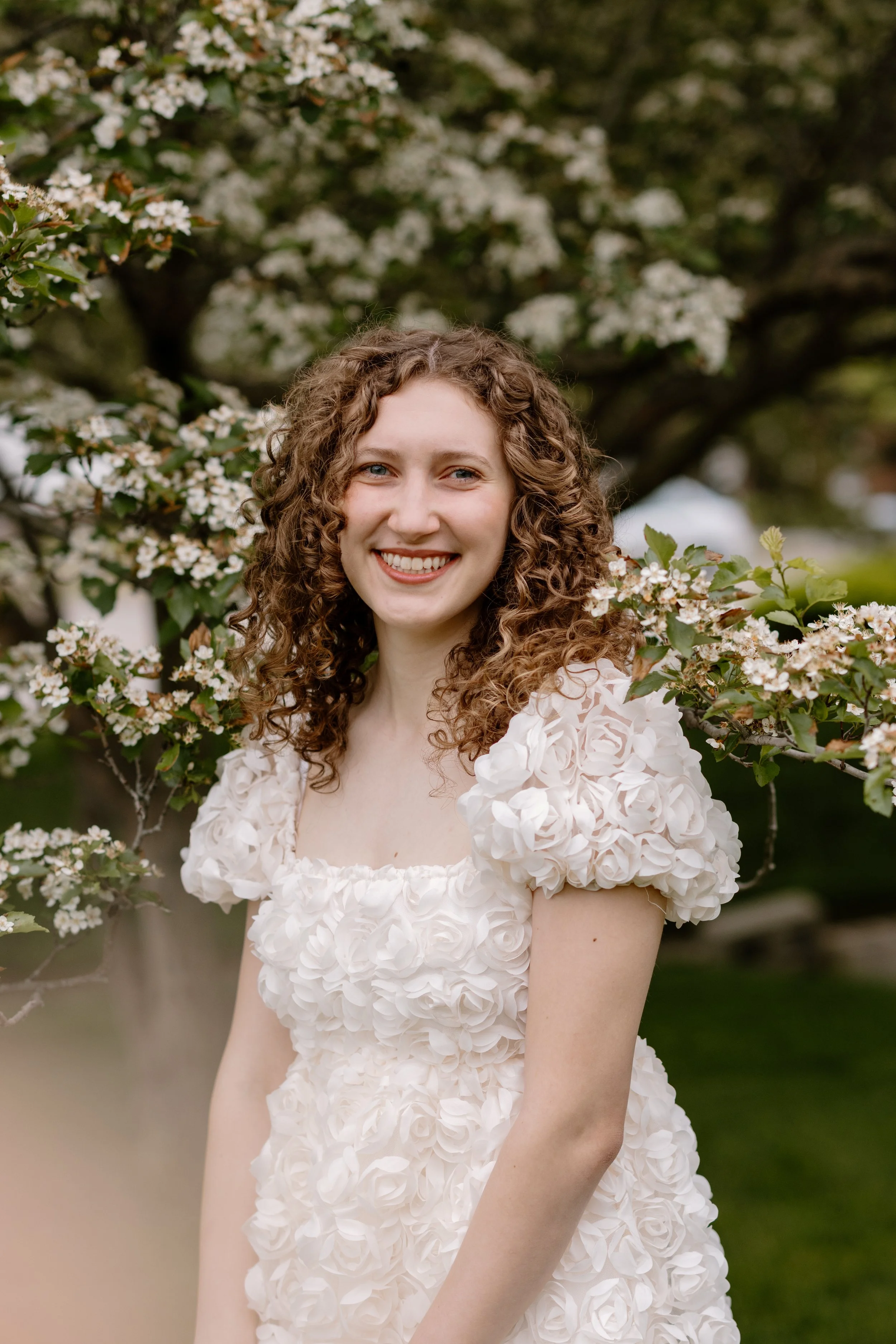 A woman with curly hair and a white floral dress smiling outdoors in front of a flowering tree.