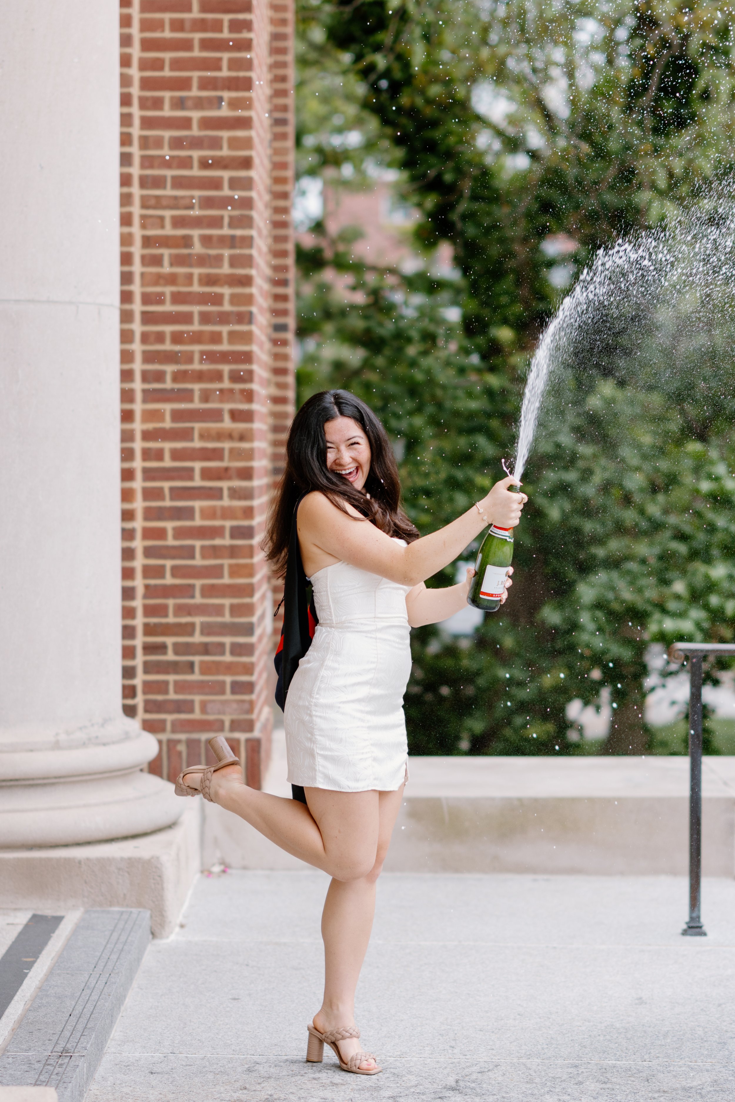 A woman in a white dress celebrating by popping open a champagne bottle with champagne spray in the air outside a brick building with trees in the background.