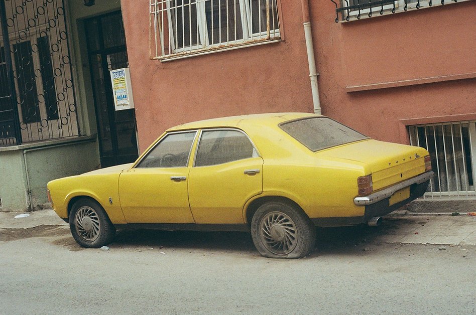 broken down, dusty yellow old ford car with flat tires in front of orange building in Izmir, Turkey. Photo by Brianna Mancini on 35mm film