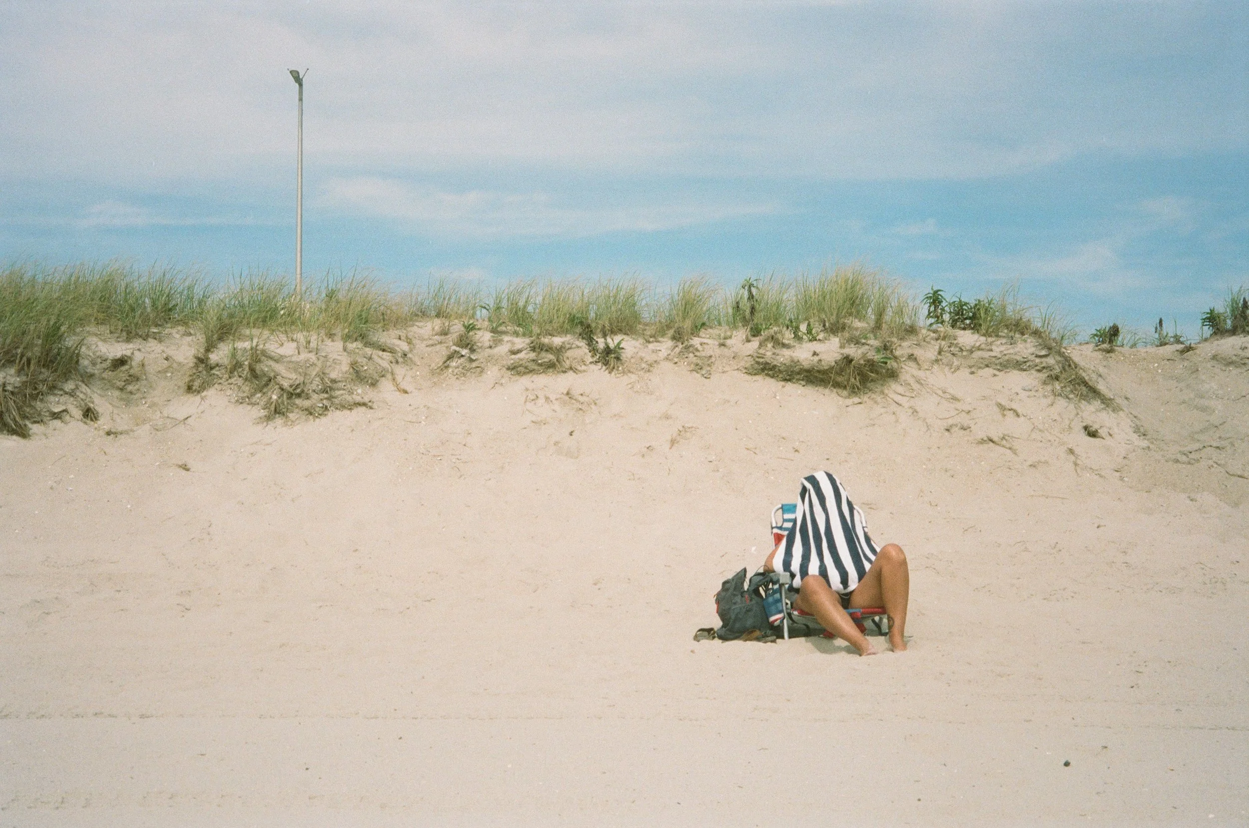 a man sitting on a beach chair in the sand with a striped towel over his head nd tall grass behind him