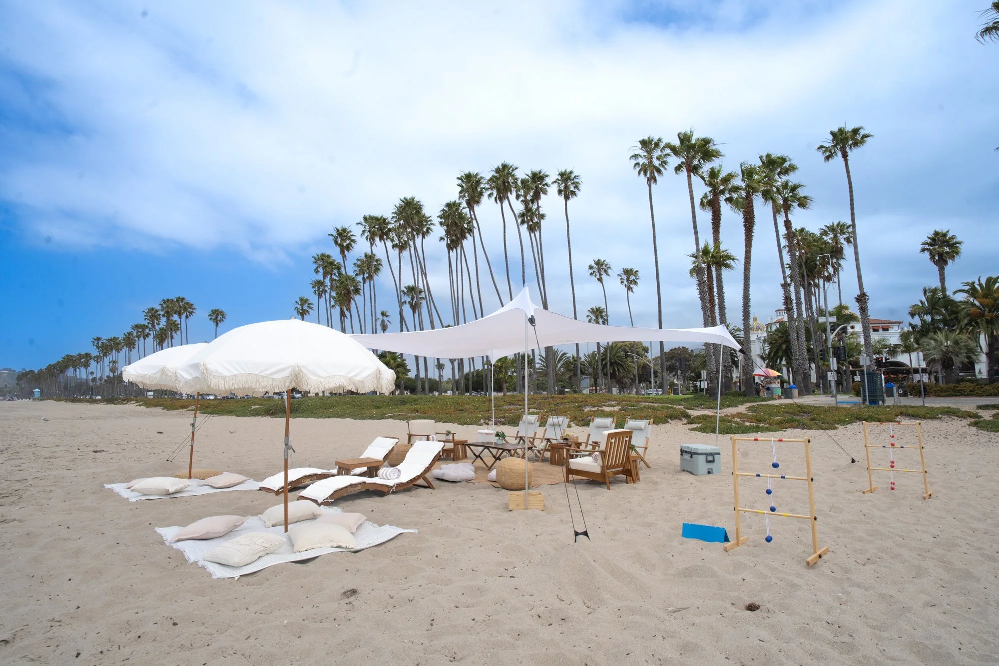 Beach setup with white umbrellas, lounge chairs, pillows, and outdoor games, with palm trees in the background and partly cloudy sky.