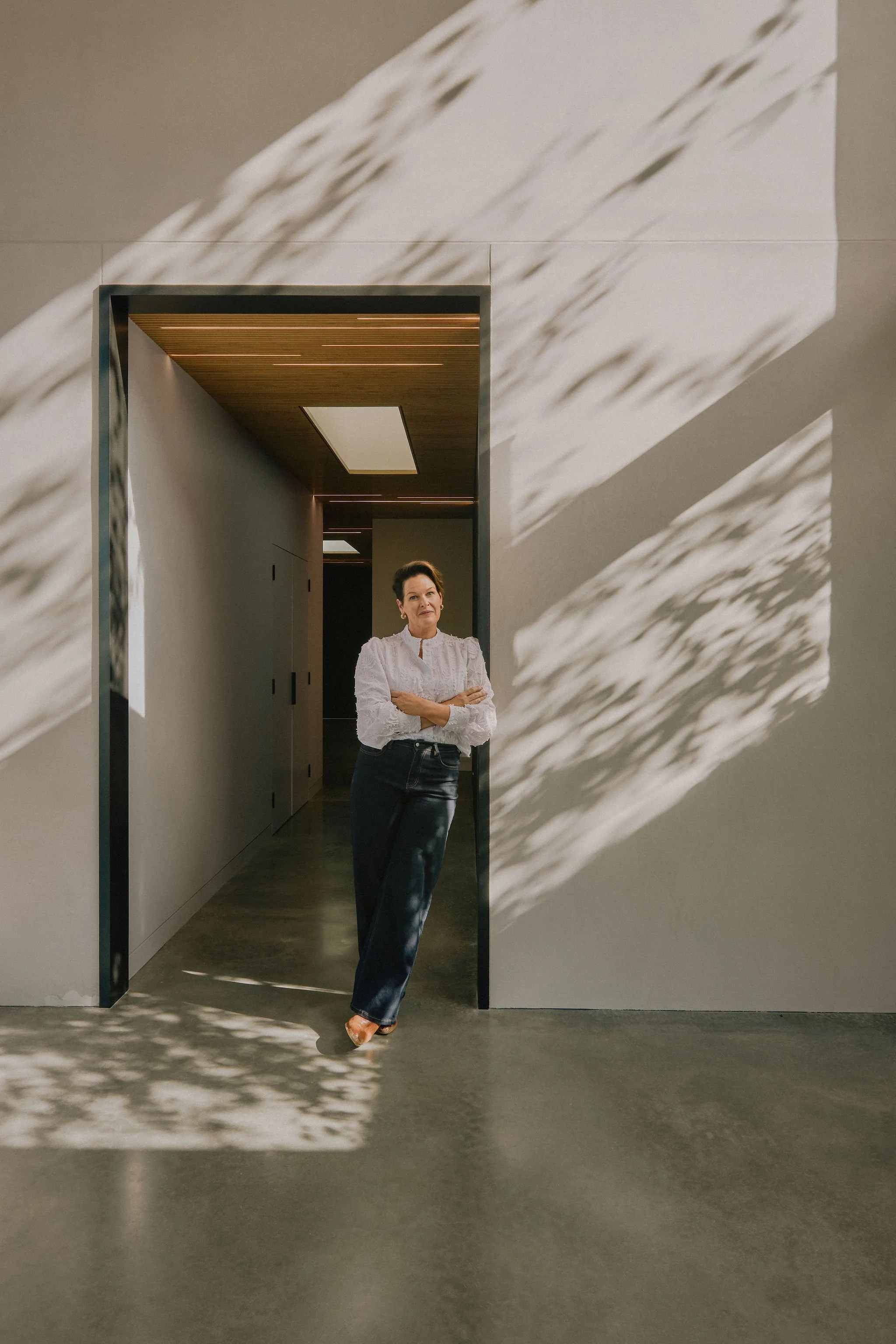 A woman with short hair in a white blouse and dark jeans stands in front of a mirror with her arms crossed, inside a minimalist room with shadows of leaves cast on the wall and floor.