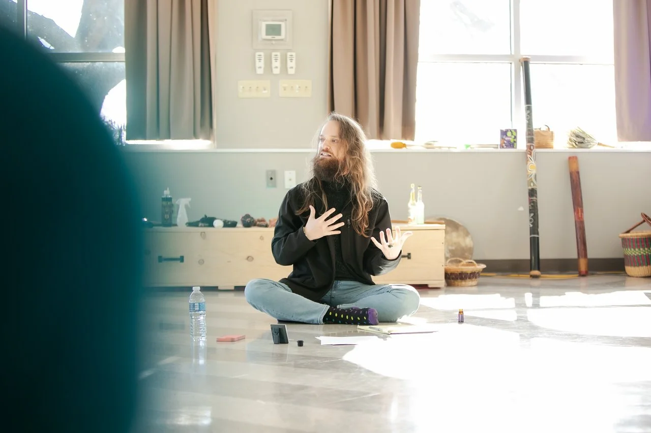 Person with long hair and a beard sitting on the floor in a room, speaking or performing, with various items around and sunlight coming through the window.